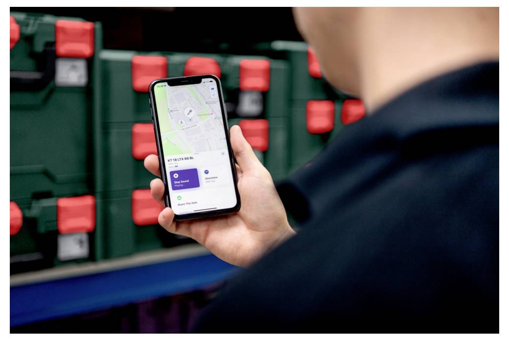 Osoba drží smartfón zobrazujúci mapu a informácie o sledovaní polohy. V pozadí sú police s zelenými a červenými krabicami.