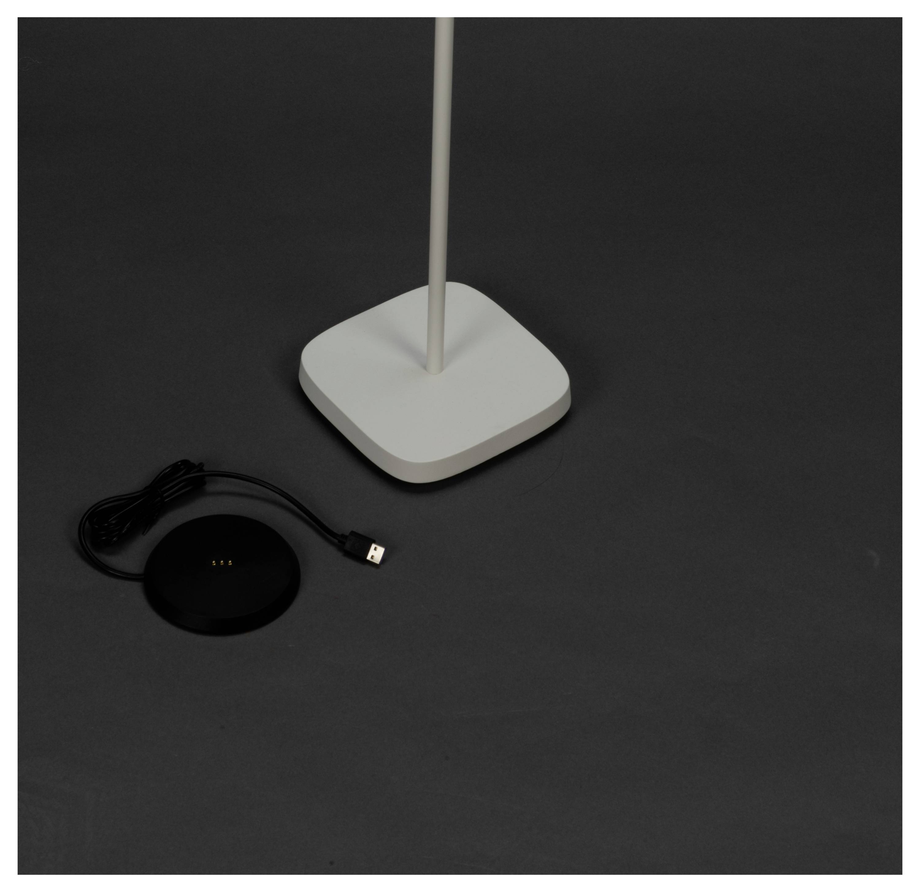 Biely štvorcový lampa stojí na tmavom povrchu, pričom vedľa je umiestnený čierny guľatý nabíjací podstavec a pripojený USB kábel.