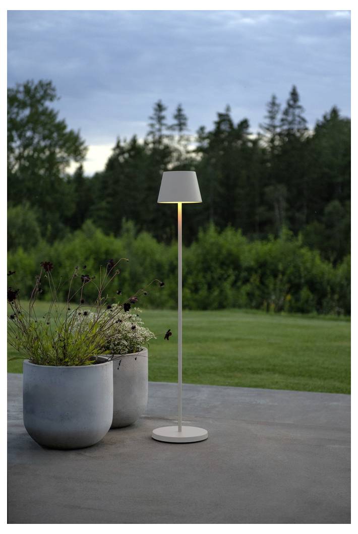 Vysoká, štíhla stojanová lampa svieti vedľa dvoch veľkých kvetináčov s rastlinami na terase, v pozadí s bohatou zelenou kulisou stromov za súmraku.