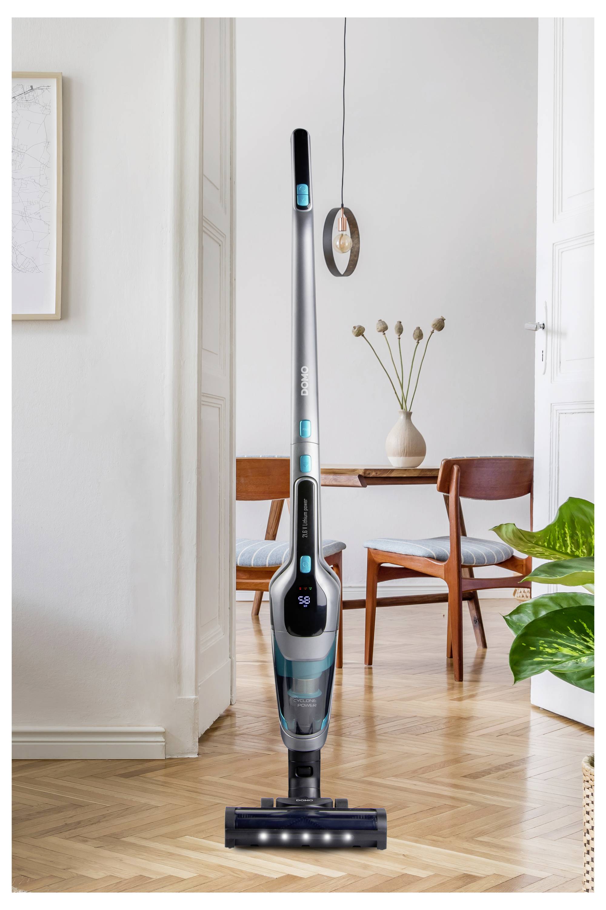 Moderný bezdrôtový vysávač stojí vzpriamene na drevenej podlahe v dobre osvetlenej obývacej izbe. V pozadí sú viditeľné jedálenský stôl a stoličky.