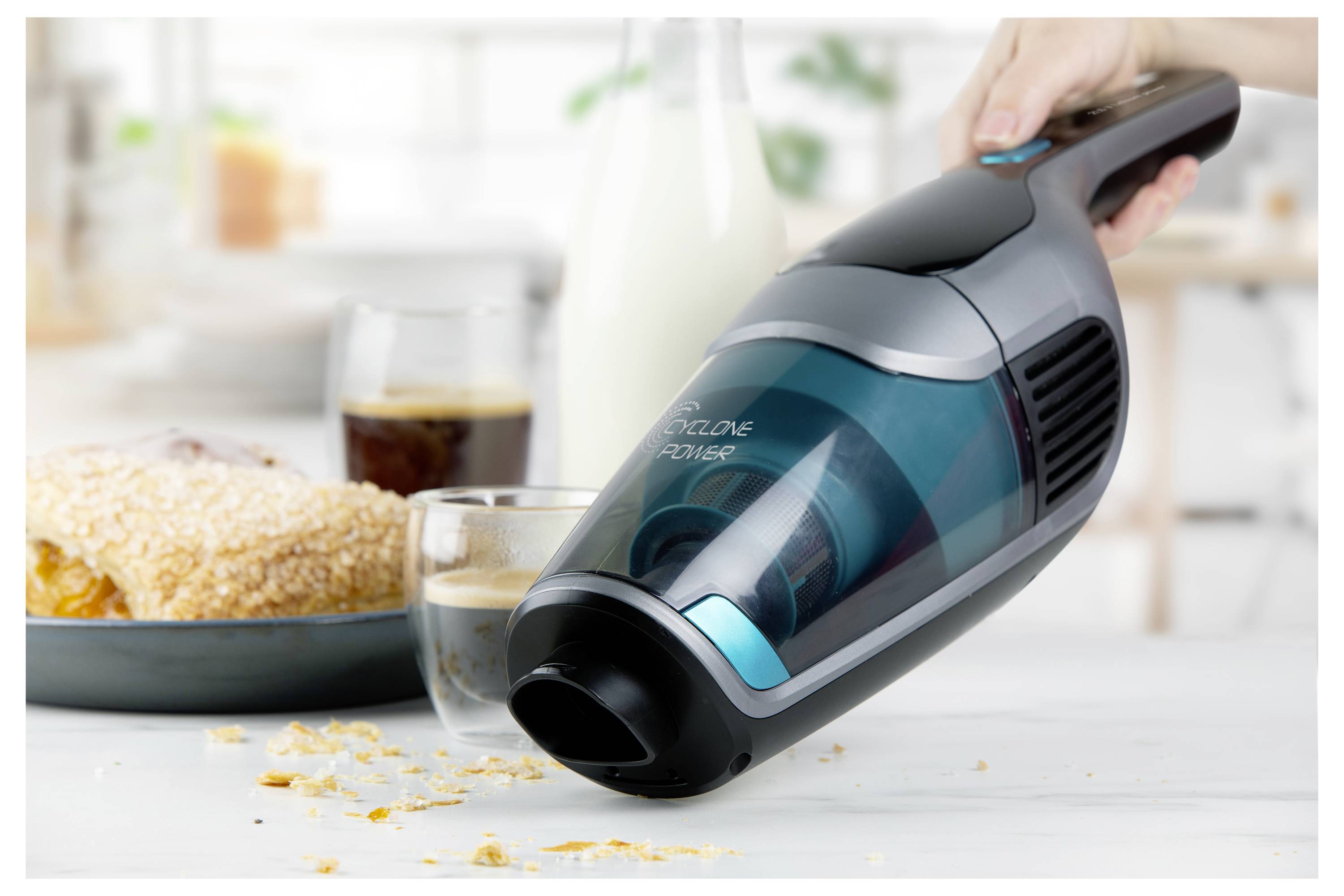 Ručný vysávač používaný na zber drobkov na bielom povrchu, v pozadí raňajkové jedlá a nápoje.