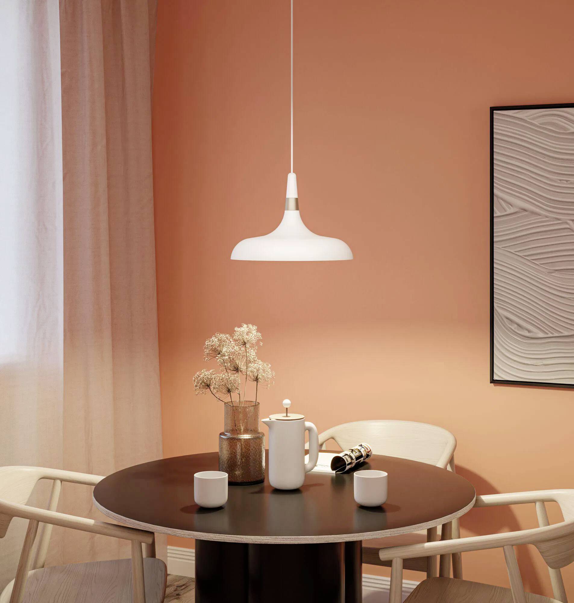 Malý okrúhly jedálenský stôl s dvoma stoličkami, kávová konvica, šálky a sušené kvety pod bielou závesnou lampou oproti oranžovej stene.