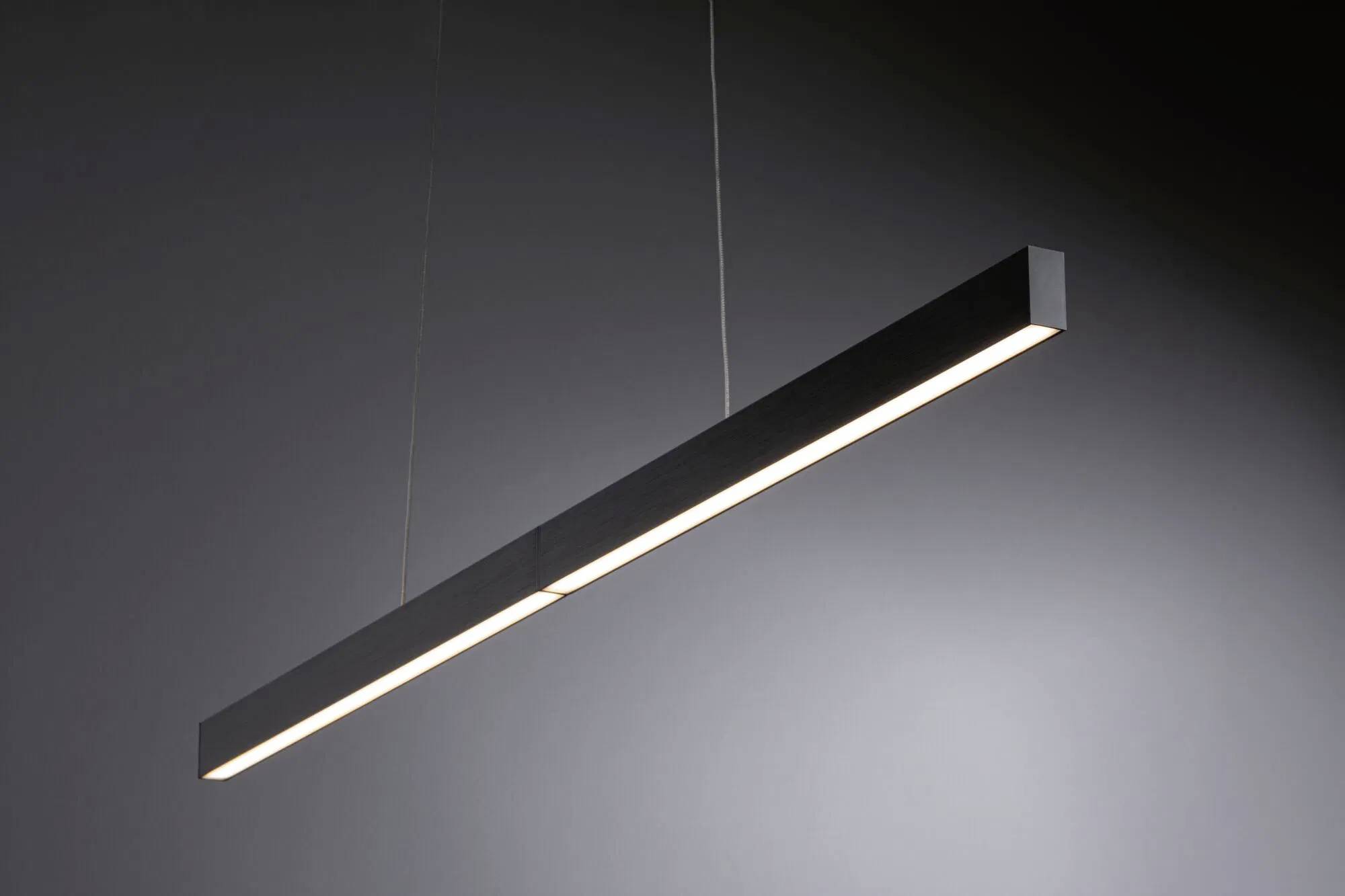 Štíhle, moderné lineárne závesné svietidlo visí pred šedým pozadím a vytvára jemný svit. Slúži ako minimalistické, elegantné osvetlenie.