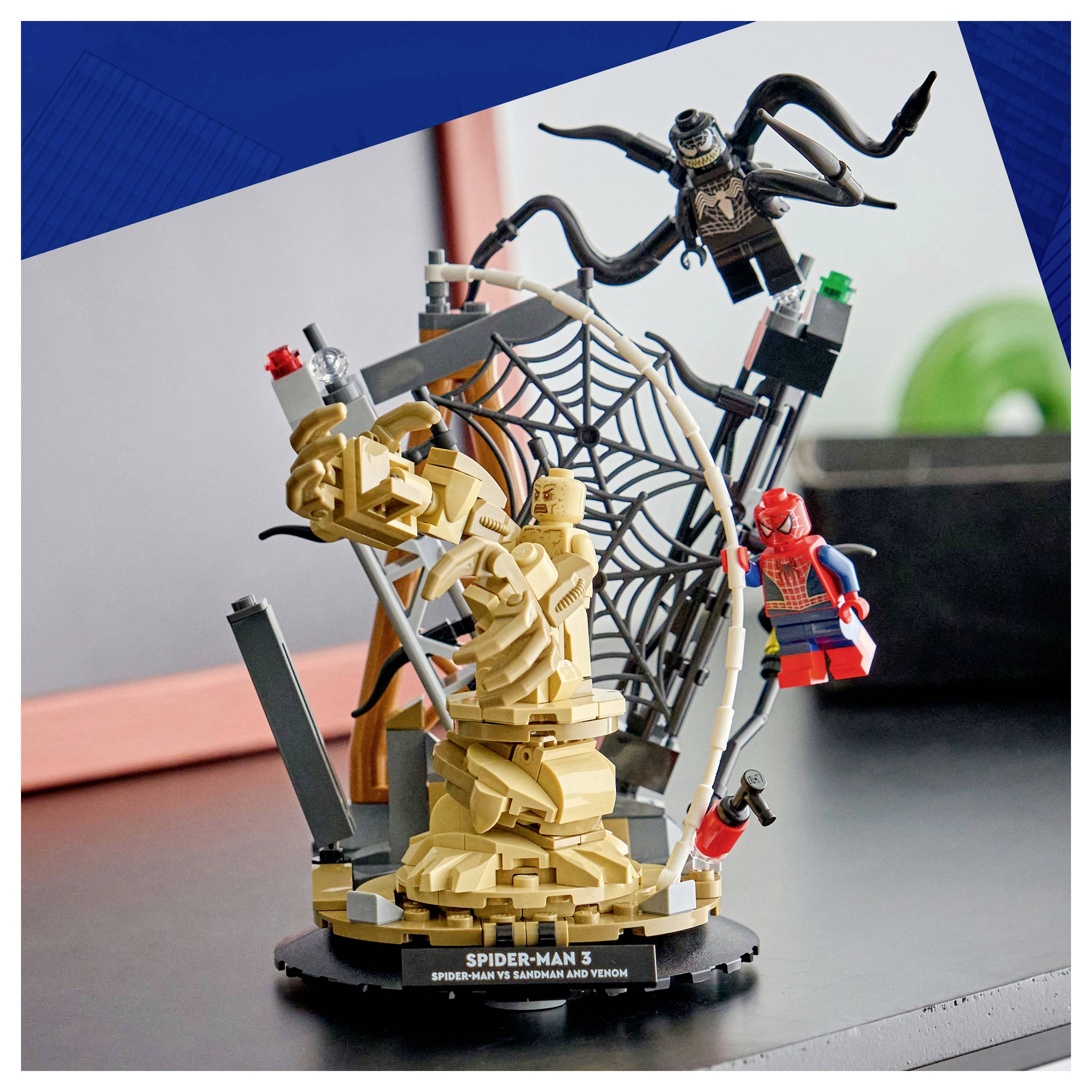 Diorama z dielikov Lego zobrazujúce Spider-Mana konfrontujúceho záporákov okolo sochársky stvárnnej figúry, obklopenej pavučinovými a lešenárskymi prvkami, umiestnené v miestnosti.