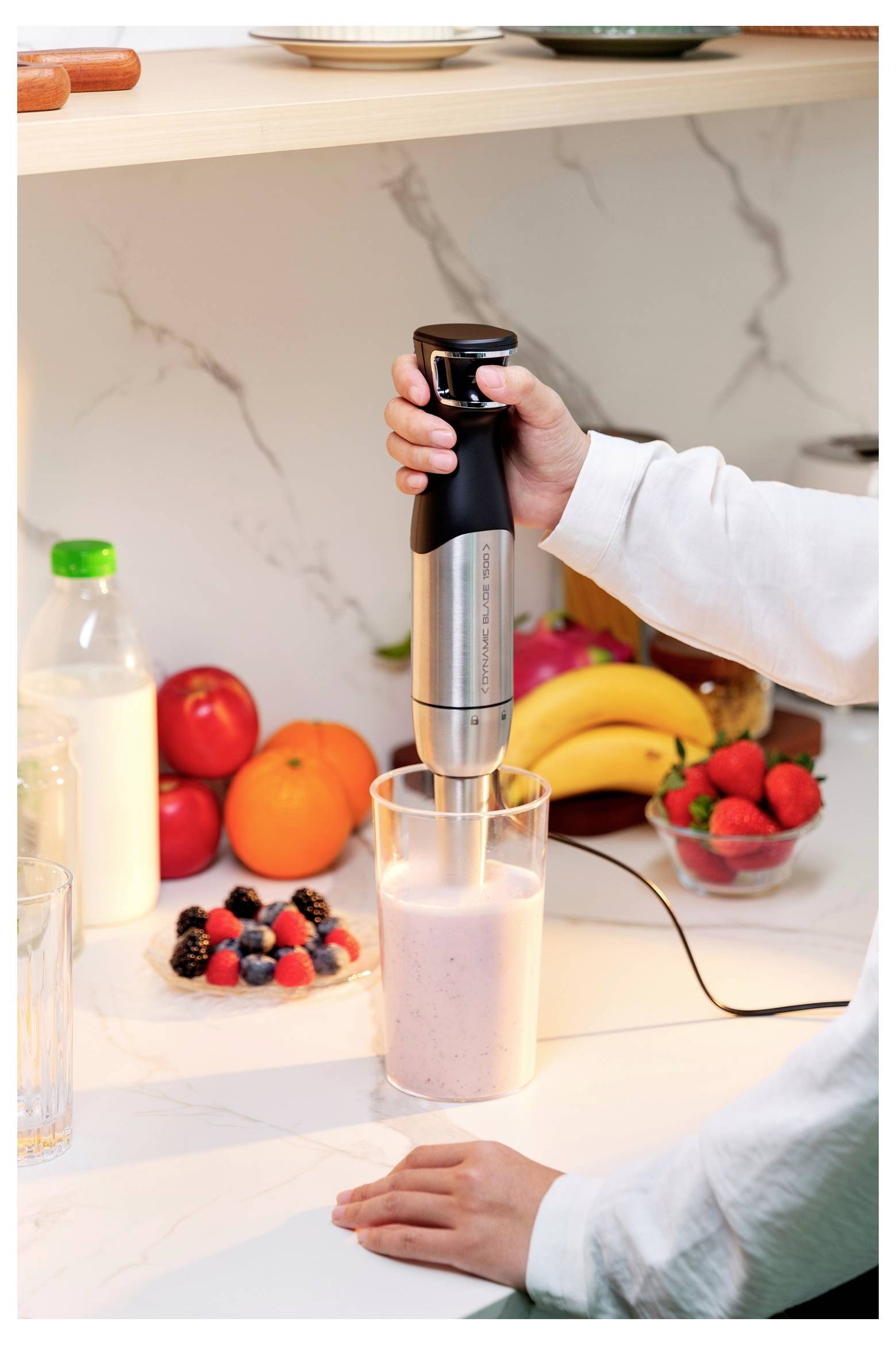 Osoba používa ponorný mixér na prípravu smoothie v kuchyni, obklopená ovocím, ako sú banány a bobuľovité plody, na kuchynskej linke.