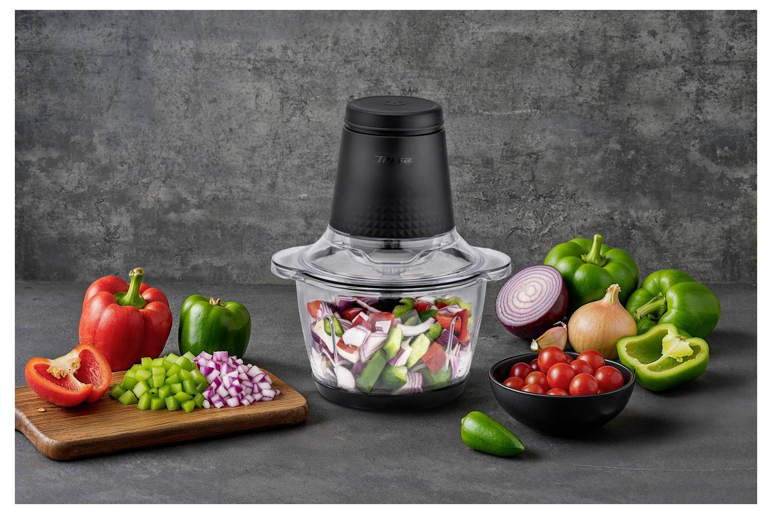 Kuchynský food processor obklopený nakrájanými zeleninou vrátane červenej a zelenej papriky, červenej cibule a cherry paradajok na pracovnej doske.