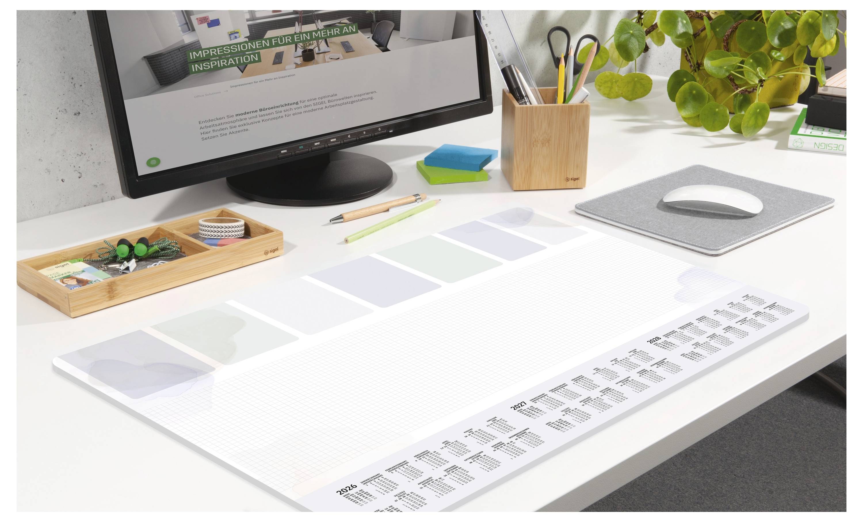 Usporiadaný stôl s kalendárom, monitorom, myšou a kancelárskymi potrebami. Zelená rastlina pridáva priestoru štipku zelene.