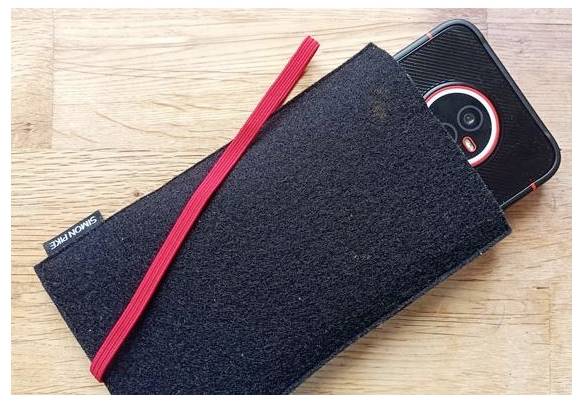 Smartphone čiastočne vyčnieva z čierneho filcového obalu, ktorý je upevnený červeným elastickým páskom, na drevenom povrchu.
