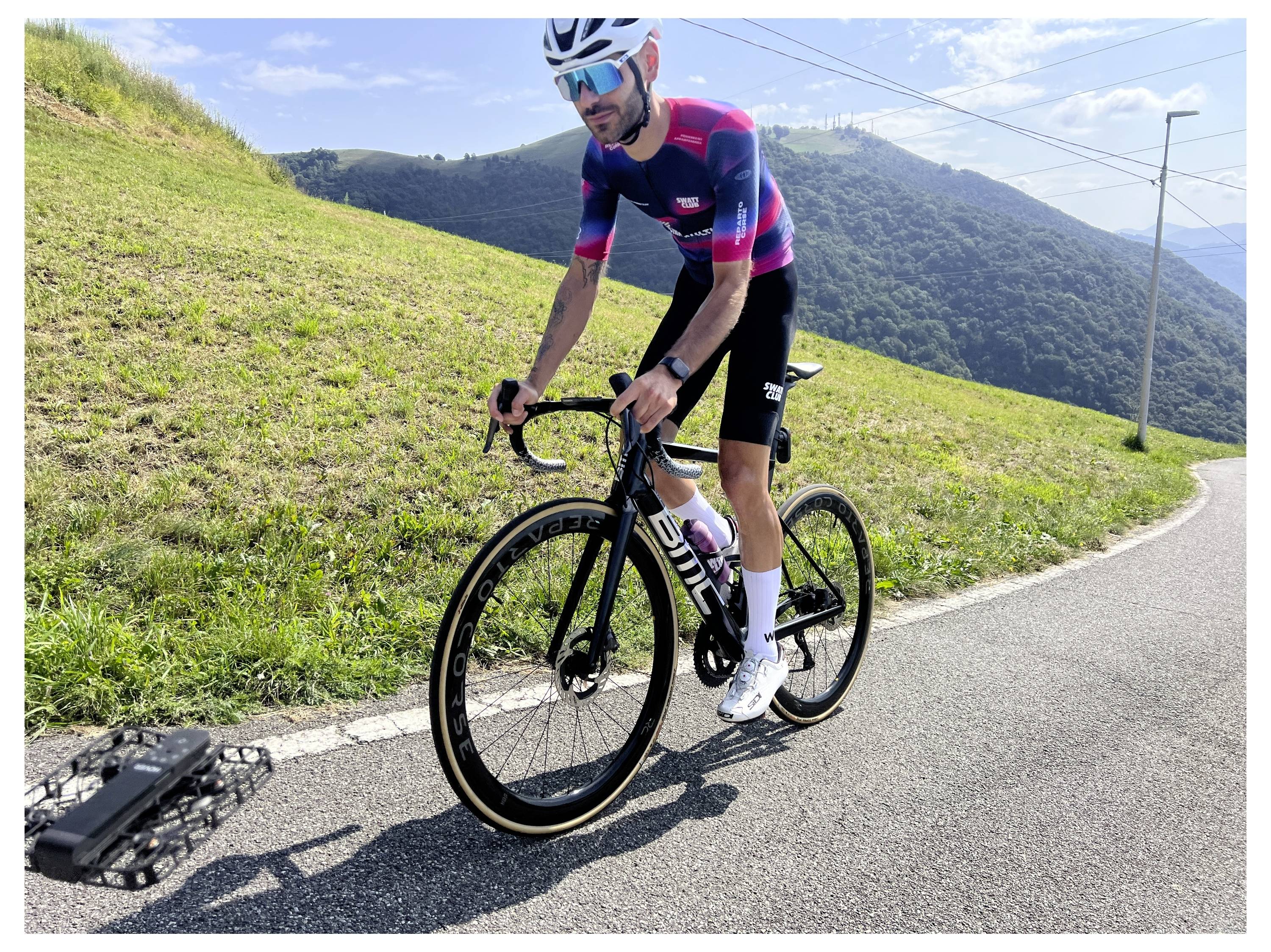Cyklista v cyklistickom oblečení jazdí na cestnom bicykli do kopca po zakrivenom cestnom úseku, v pozadí so zelenými kopcami a jasnou modrou oblohou.