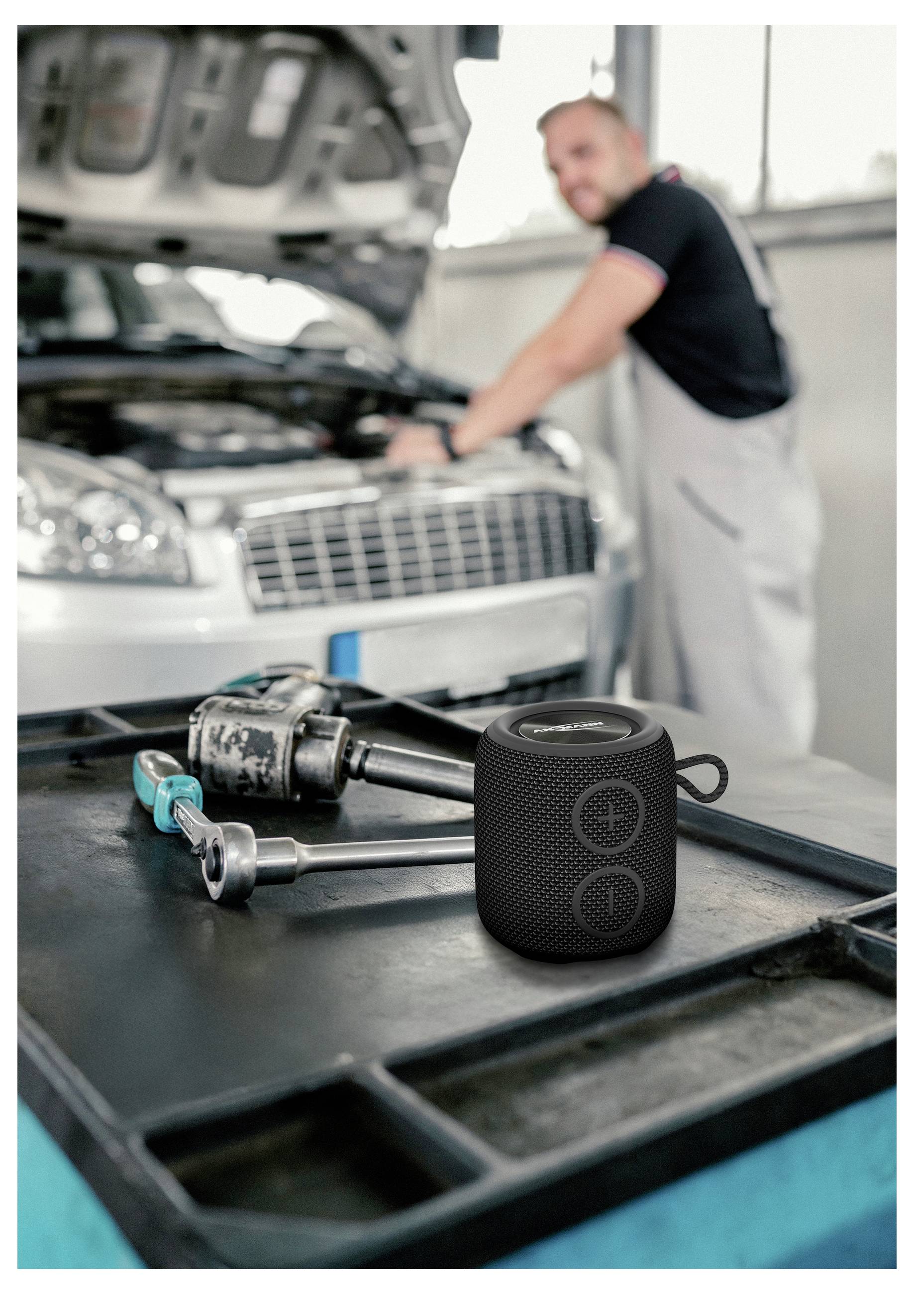 Prenosný reproduktor na pracovnom stole v autoservise, pričom mechanik pracuje pod otvorenou kapotou automobilu v pozadí.