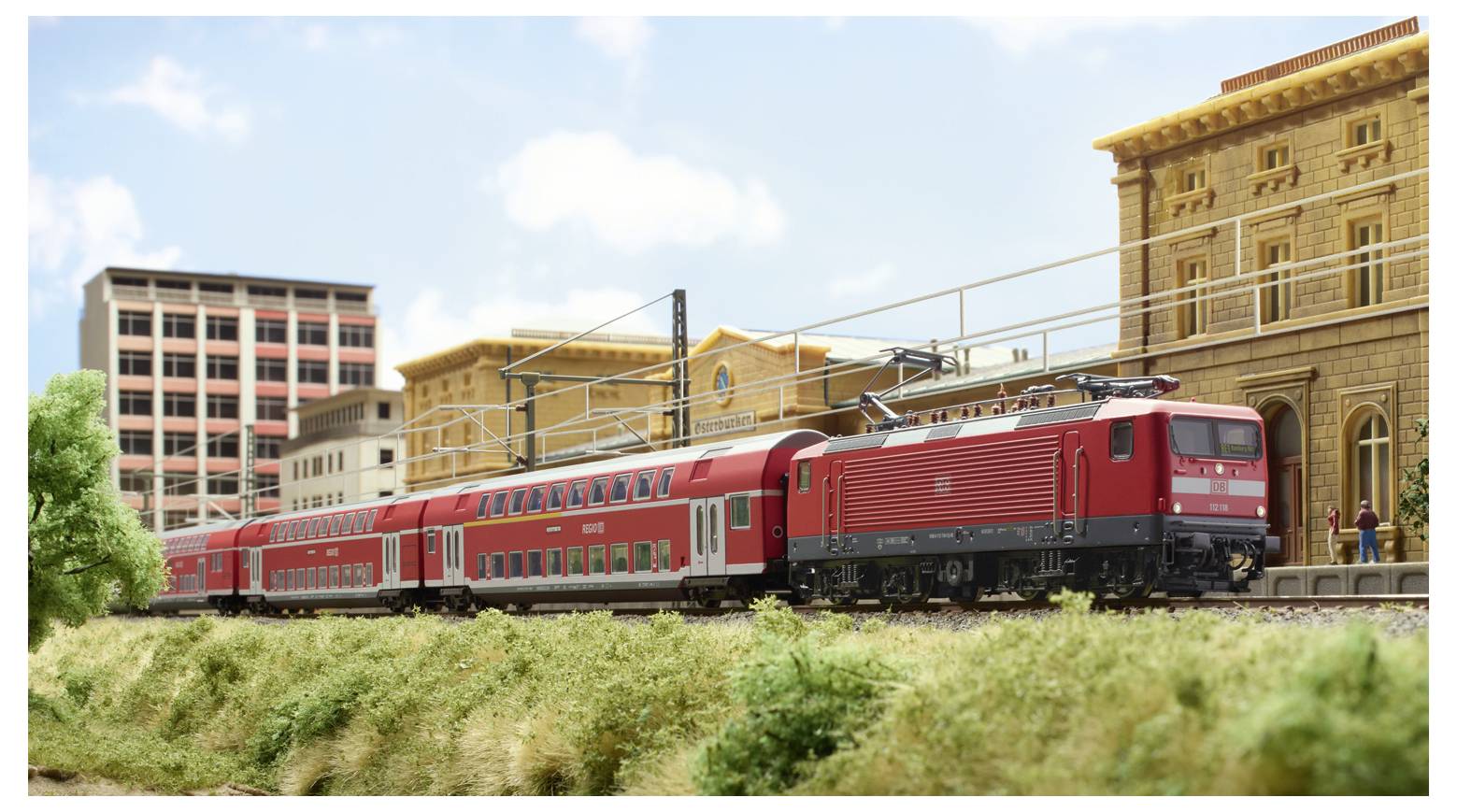 Červený vlak prechádza modelovým mestom s detailne spracovanými budovami a zeleňou, čím prezentuje železničnú scénu s mestskou architektúrou.