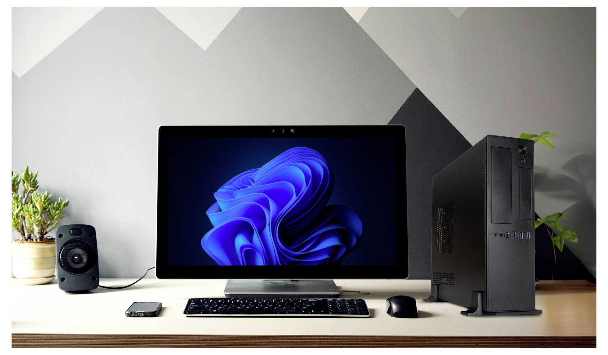 Upravený pracovný stôl s počítačovým monitorom zobrazujúcim modrý abstraktný dizajn, klávesnicou, myšou, smartfónom, reproduktorom a stolným počítačom.