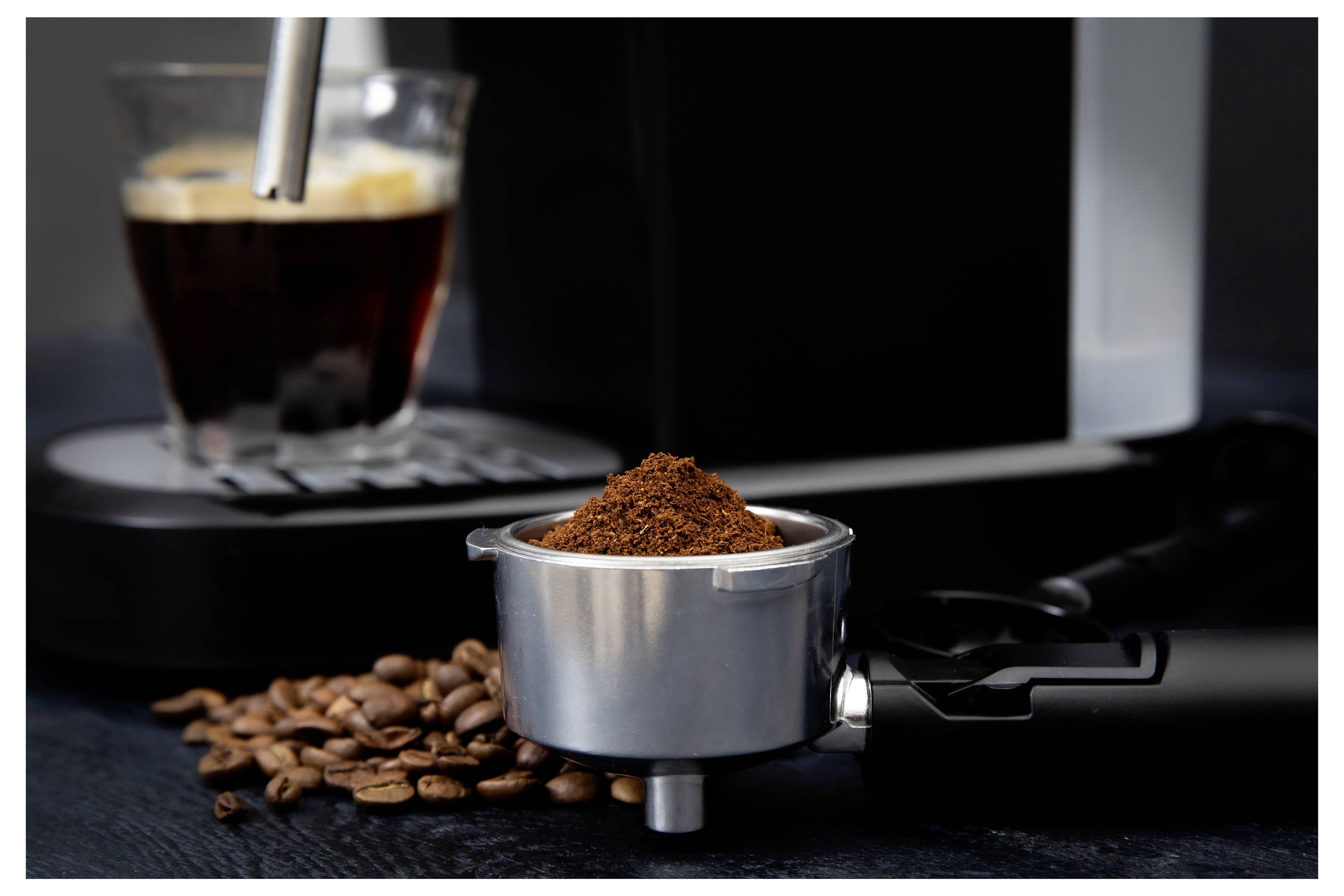Portafiltier naplnený mletou kávou je položený vedľa roztrúsených kávových zŕn a espresso stroja, pričom v pozadí je pripravené espresso.