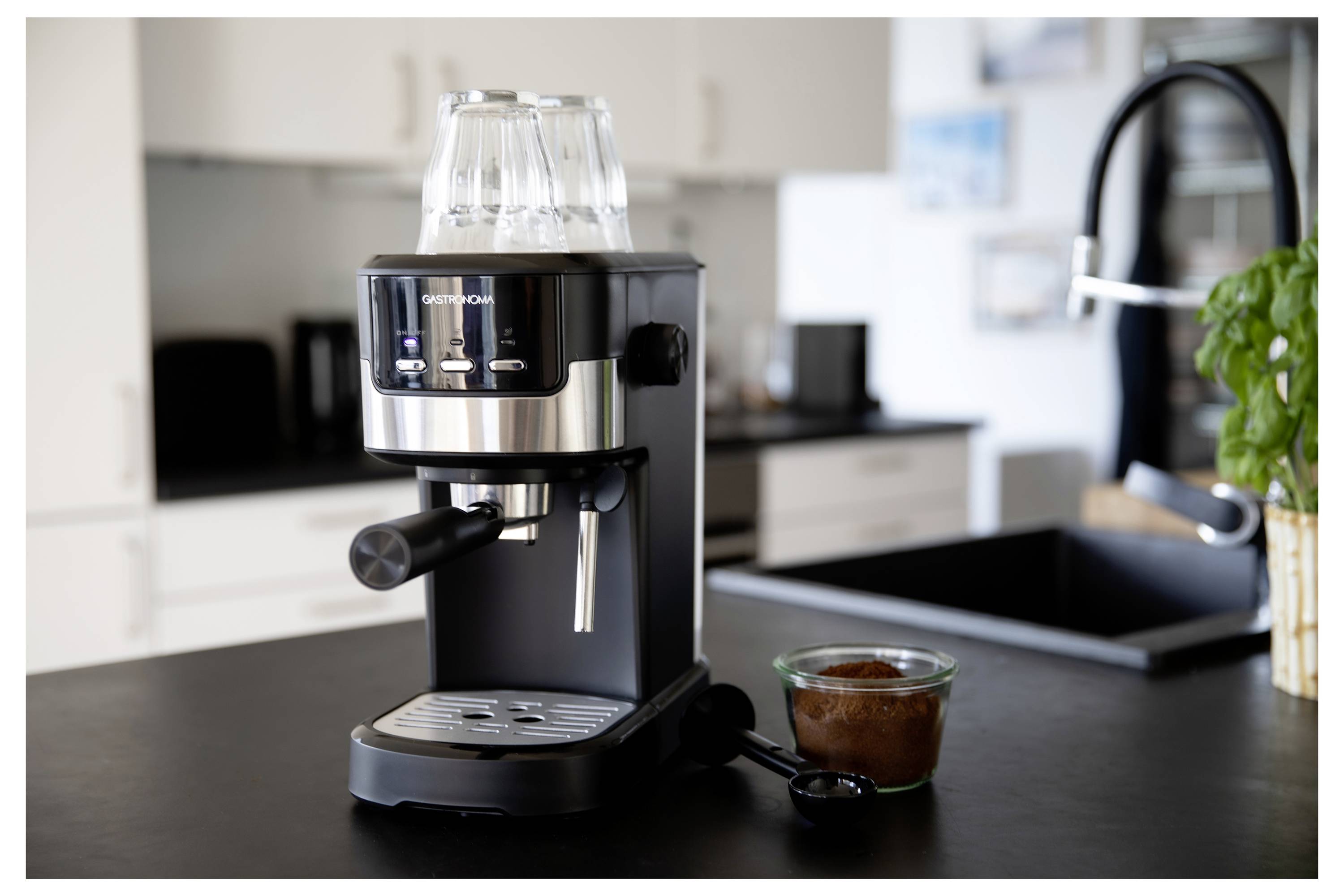 Čierna a nehrdzavejúca oceľová espresso kávovar na kuchynskej linke s šálkou mletej kávy a naukladanými šálkami na vrchu stroja.