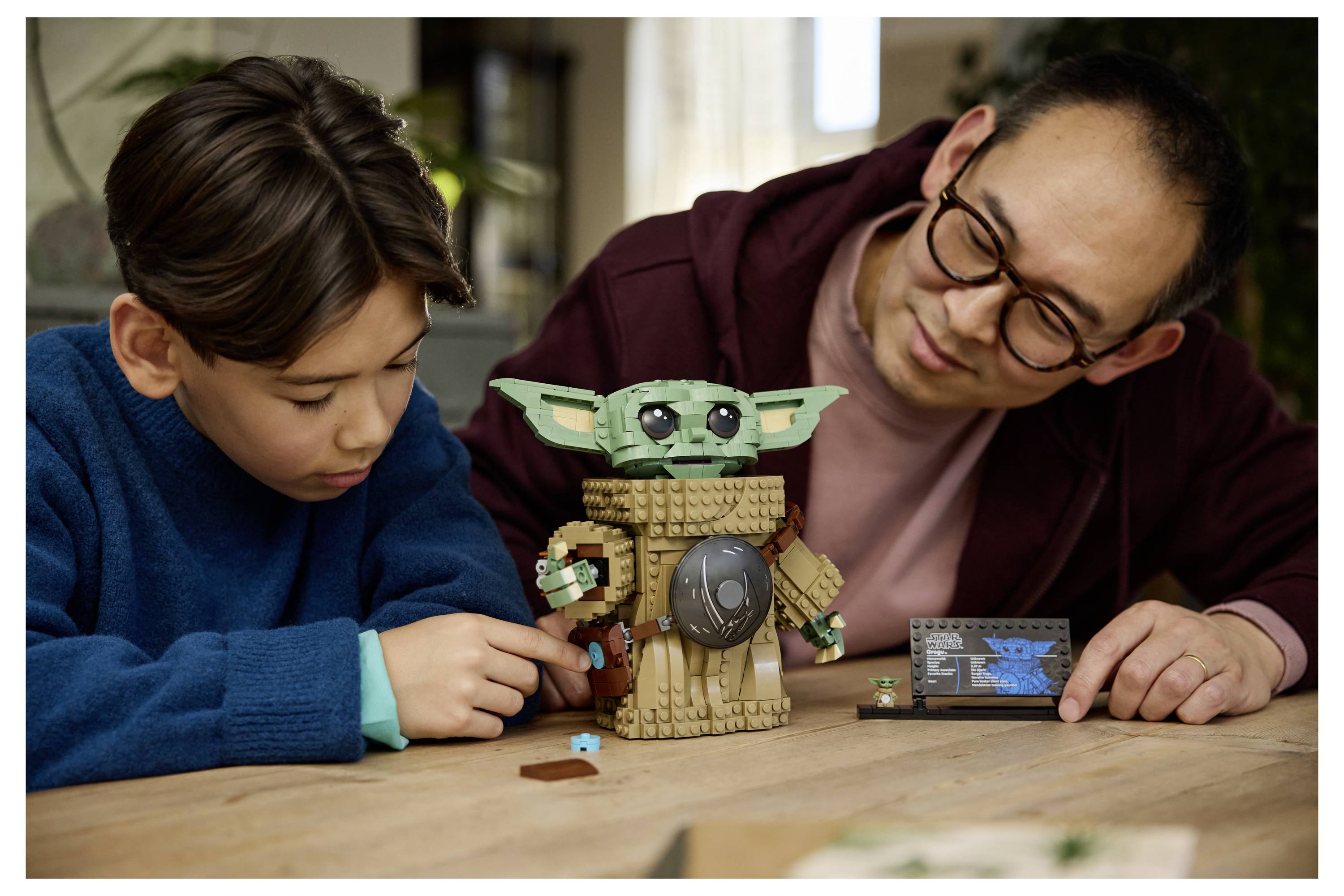 Mladý chlapec a dospelý človek montujú sadu LEGO „Star Wars: The Mandalorian" s figúrkou Grogu na stole v útulnej miestnosti.