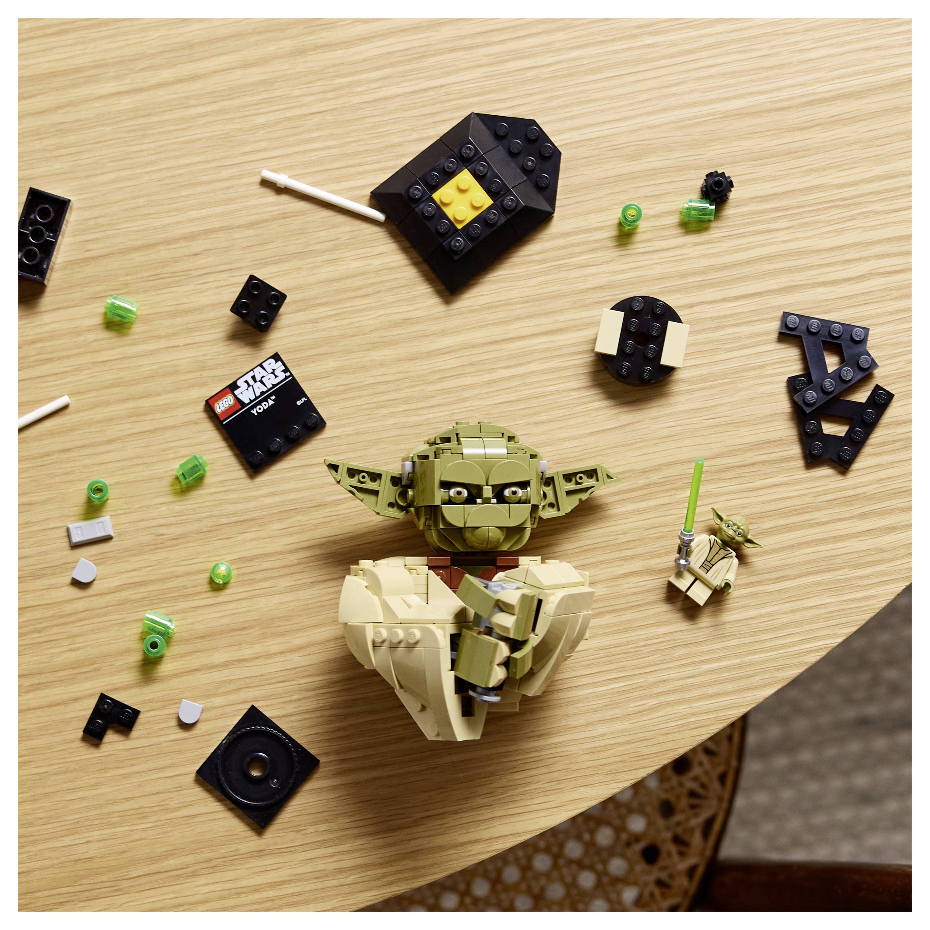 Čiastočne postavená LEGO figúrka Yodu na drevenom stole, obklopená roztrúsenými kockami LEGO a mini LEGO Yodom so svetelným mečom.