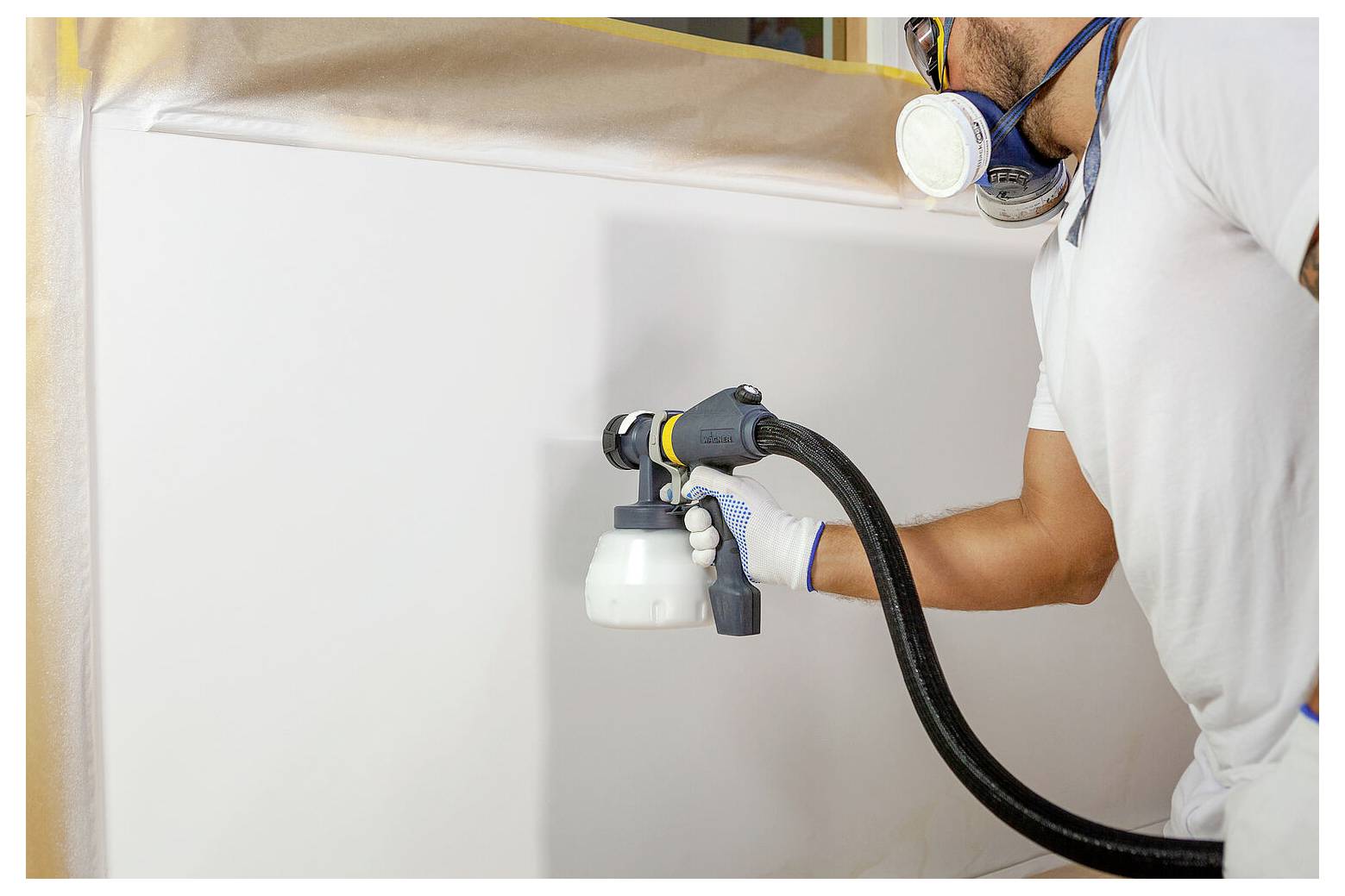 Osoba striekajúca veľkú plochu striekacou pištoľou, ktorá má na sebe respirátor a rukavice na ochranu.