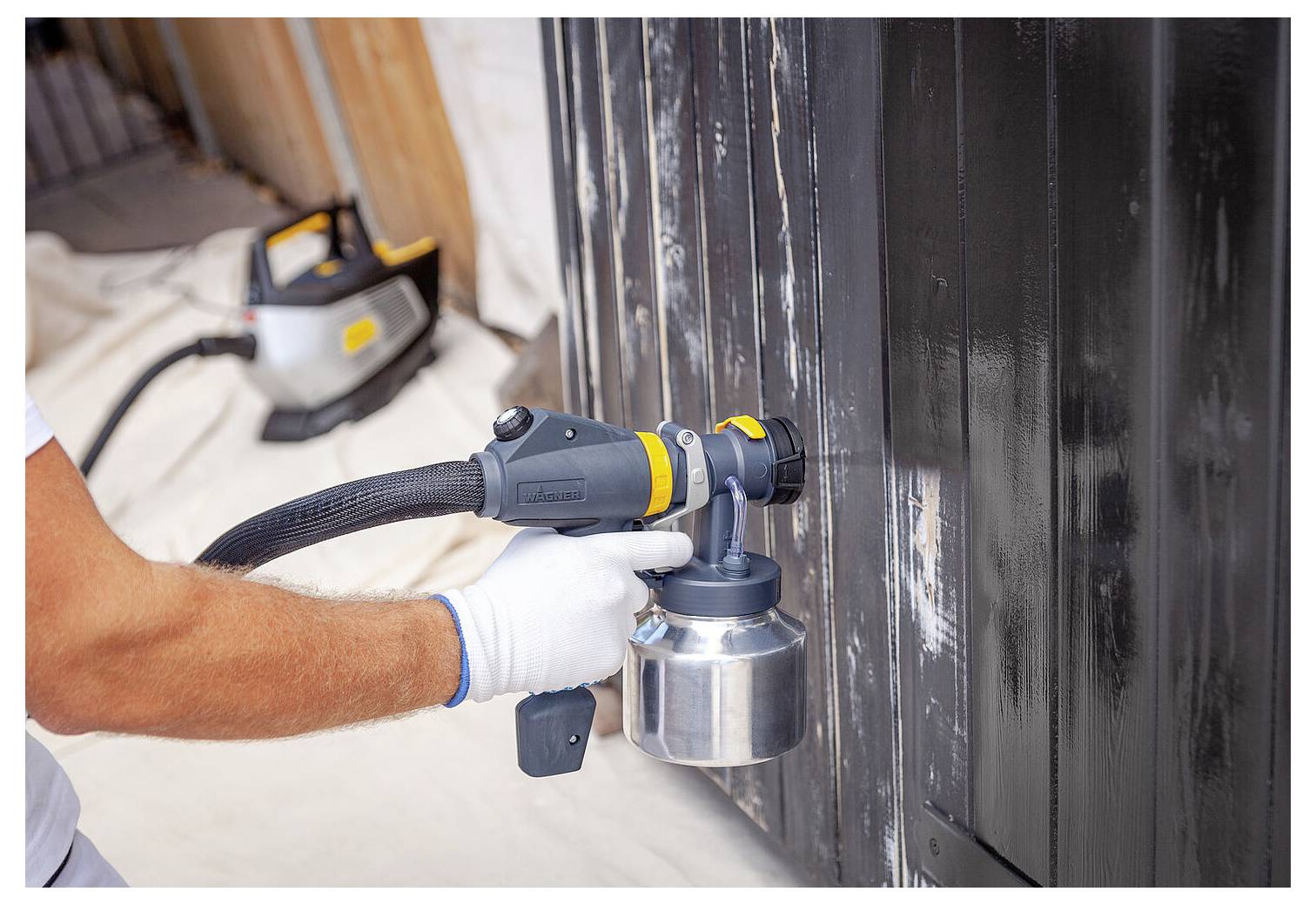 'Osoba používa striekaciu pištoľ na nanášanie čiernej farby na drevený povrch, nachádza sa v otvorenom priestore s ochrannými pokrývkami na podlahe.'