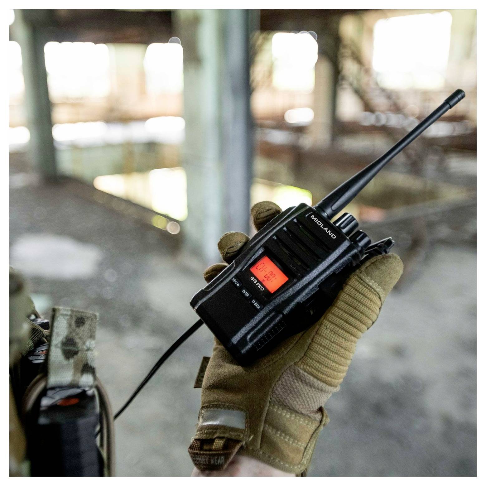 Osoba v taktických rukaviciach drží vysielačku (walkie-talkie) v interiéri, čím zdôrazňuje komunikačné zariadenie v priemyselnom prostredí.