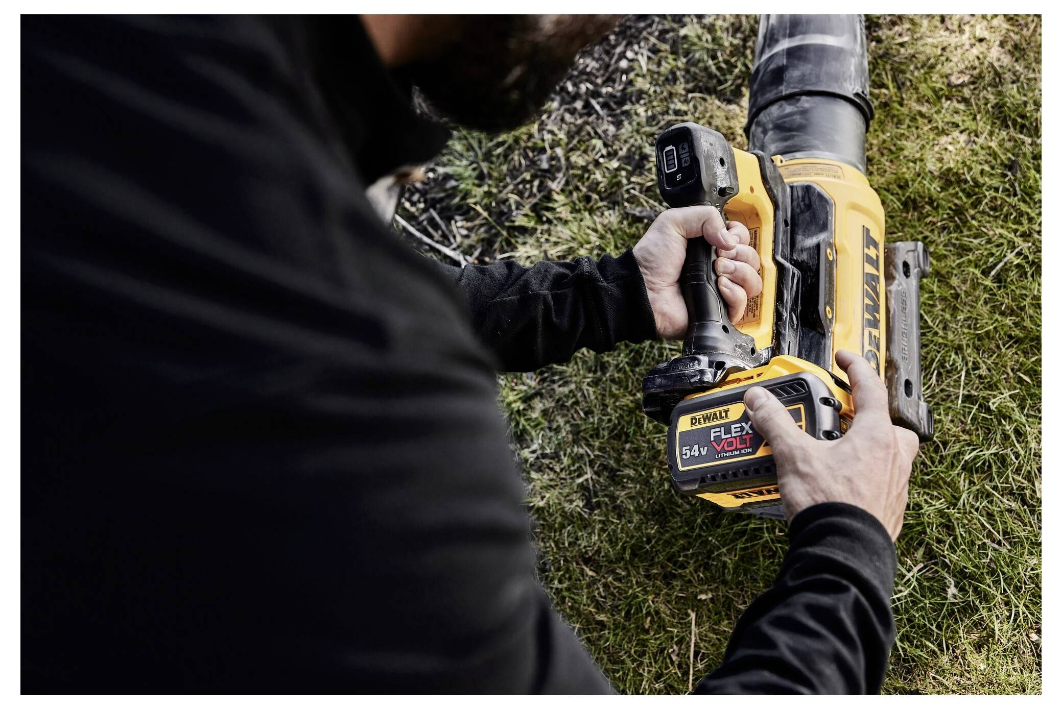 Osoba používa čierno-žltý elektrický nástroj značky „DeWalt Flex Volt 54V" na trávnatej ploche a zameriava sa na jeho nastavenie.
