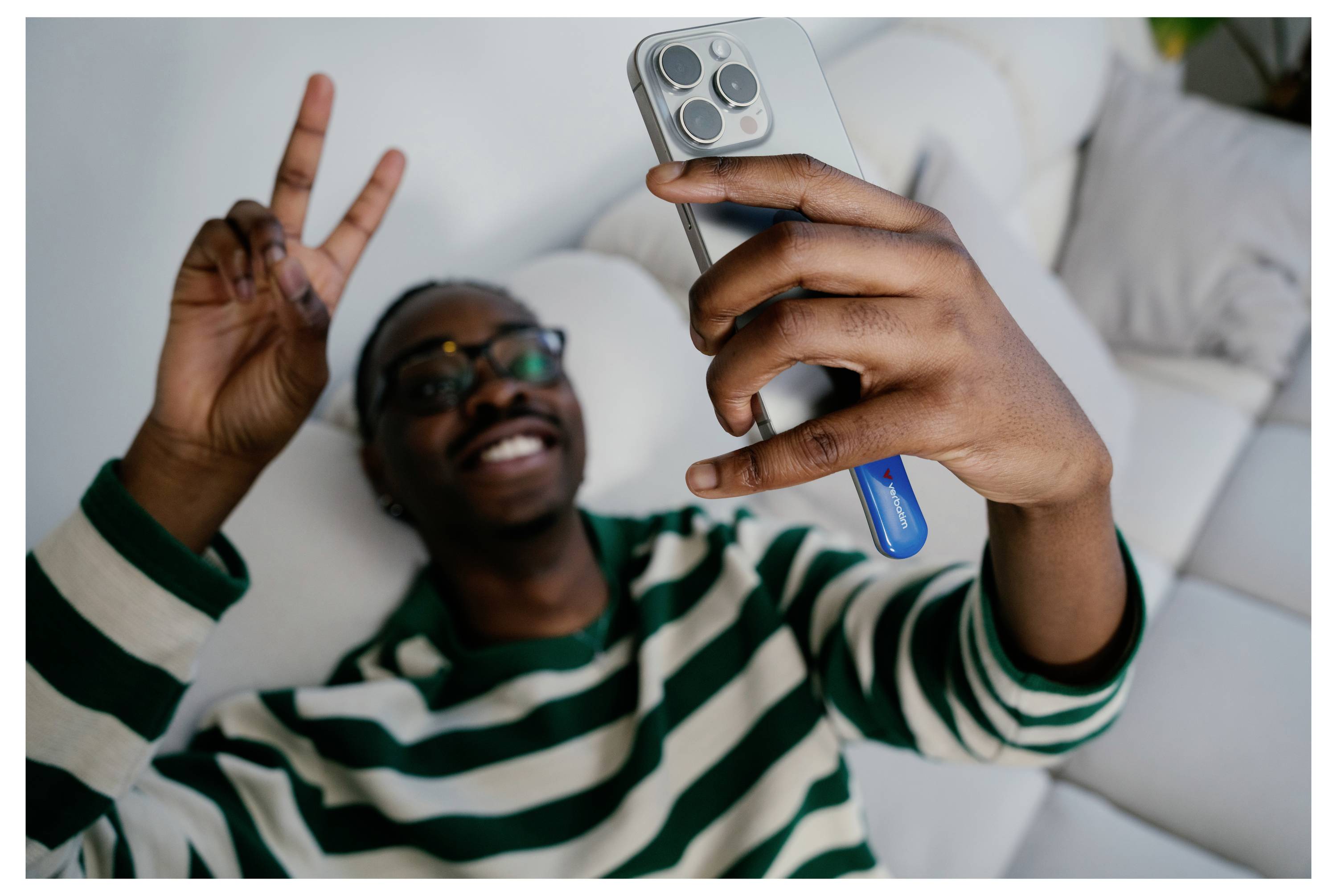 Úsmevný muž v pruhovanom svetre si robí selfie a ukazuje piace znamenie zatiaľ čo leží na posteli.