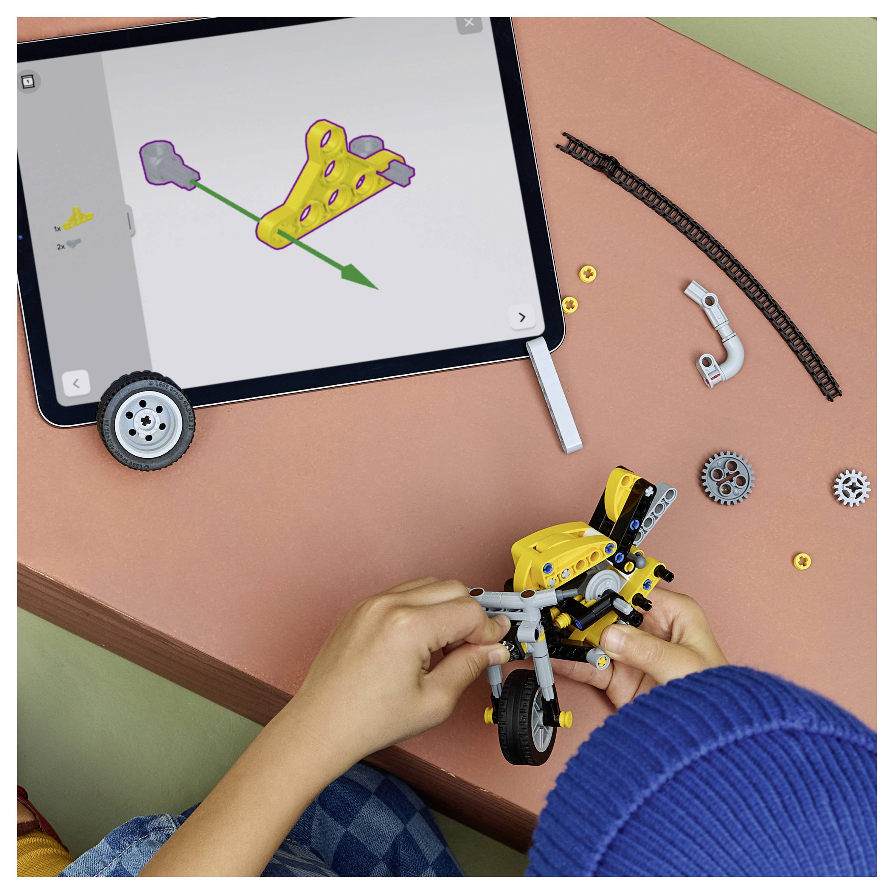 Dieťa skladá žltú robotickú hračku motorky na stole a sleduje pokyny zobrazené na tablete.