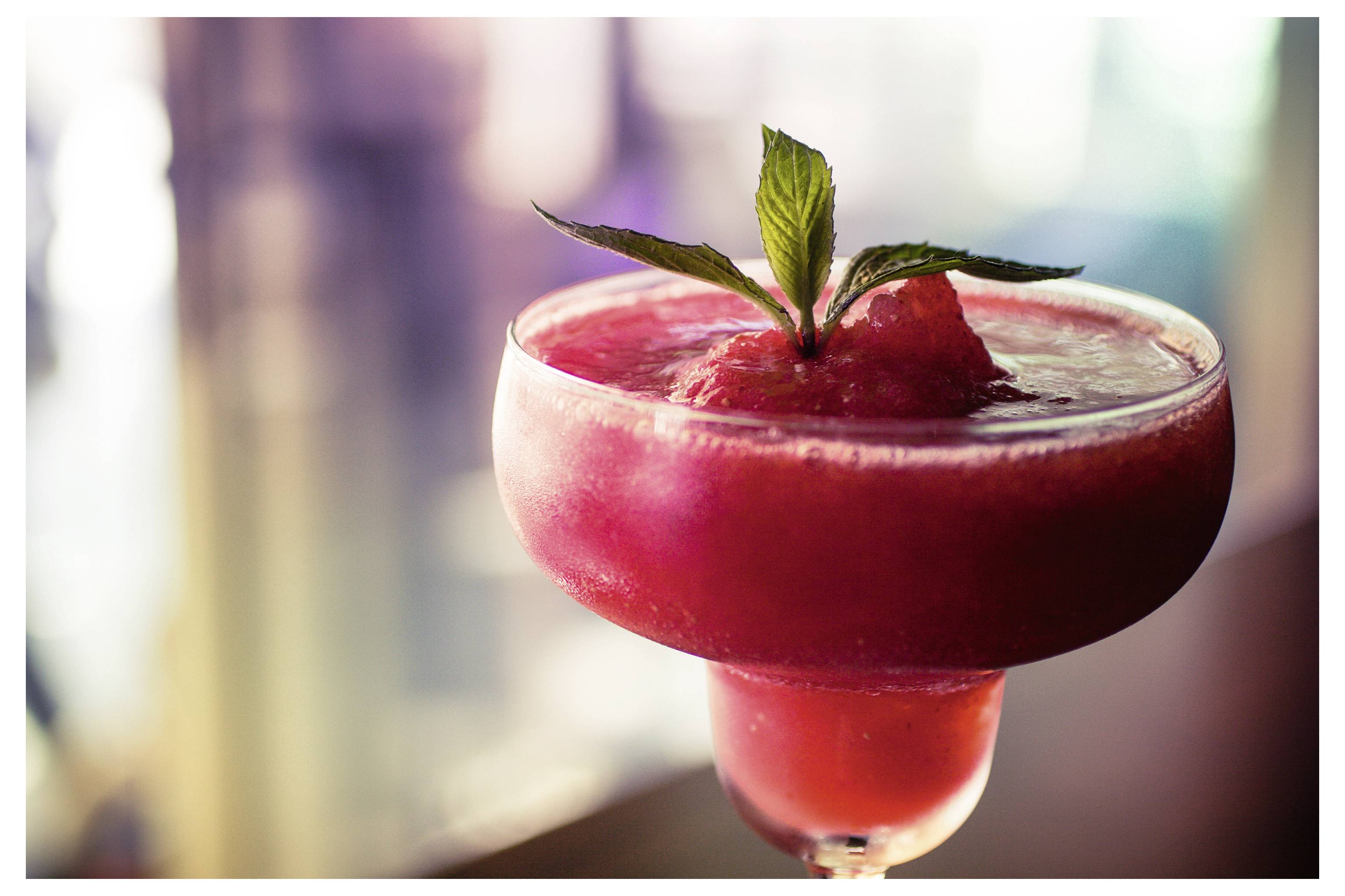 Mrazivý červený koktejl v pohári, ozdobený listami mäty, na pozadí rozmazaného prostredí.