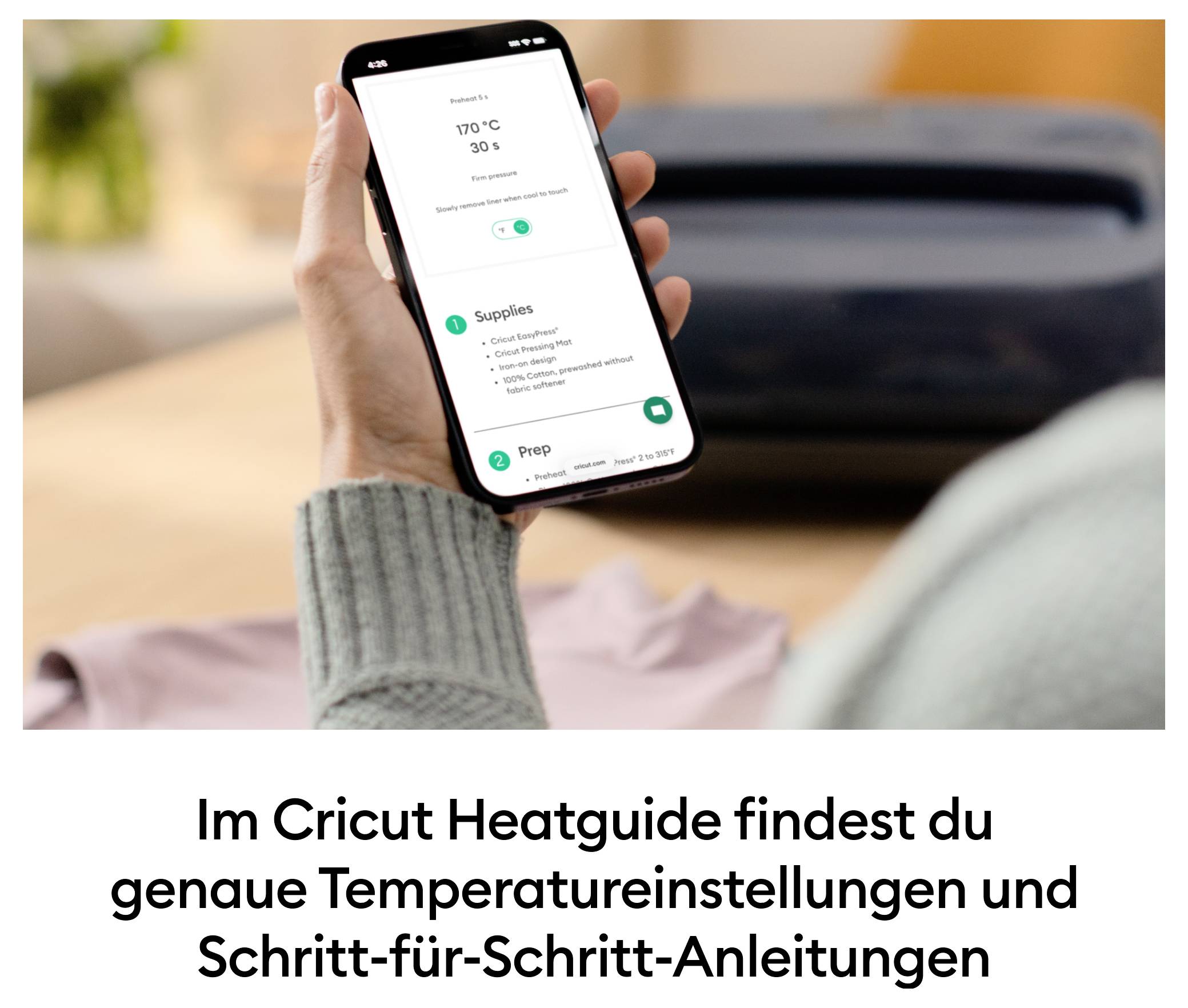 Osoba drží smartfón s aplikáciou Cricut Heatguide, ktorá zobrazuje teploty a pokyny pre projekty. Na stole leží zariadenie Cricut.