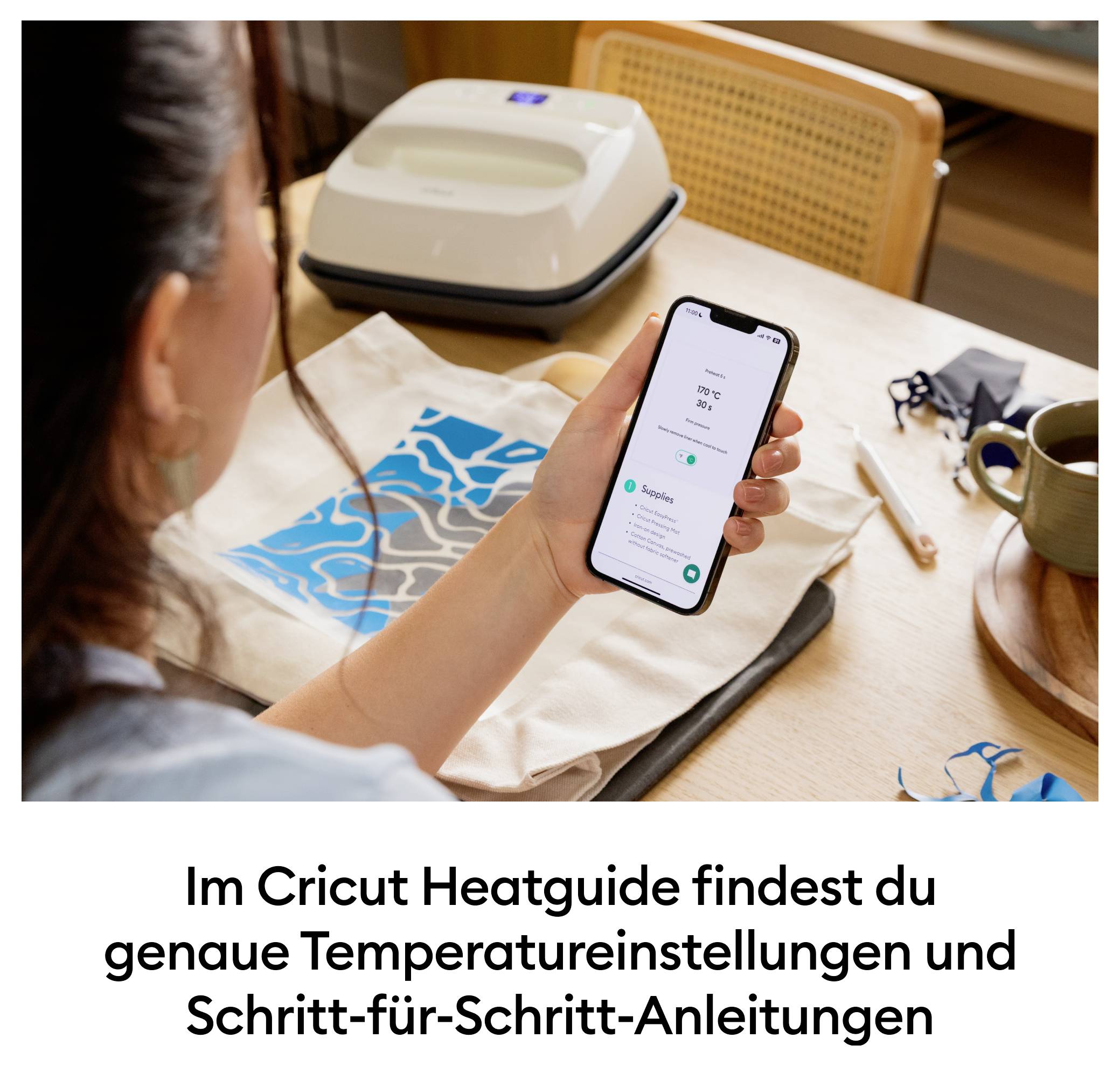 Osoba drží smartfón s otvorenou aplikáciou Cricut Heatguide. V pozadí je viditeľný prenosný lis a projekt na textil.