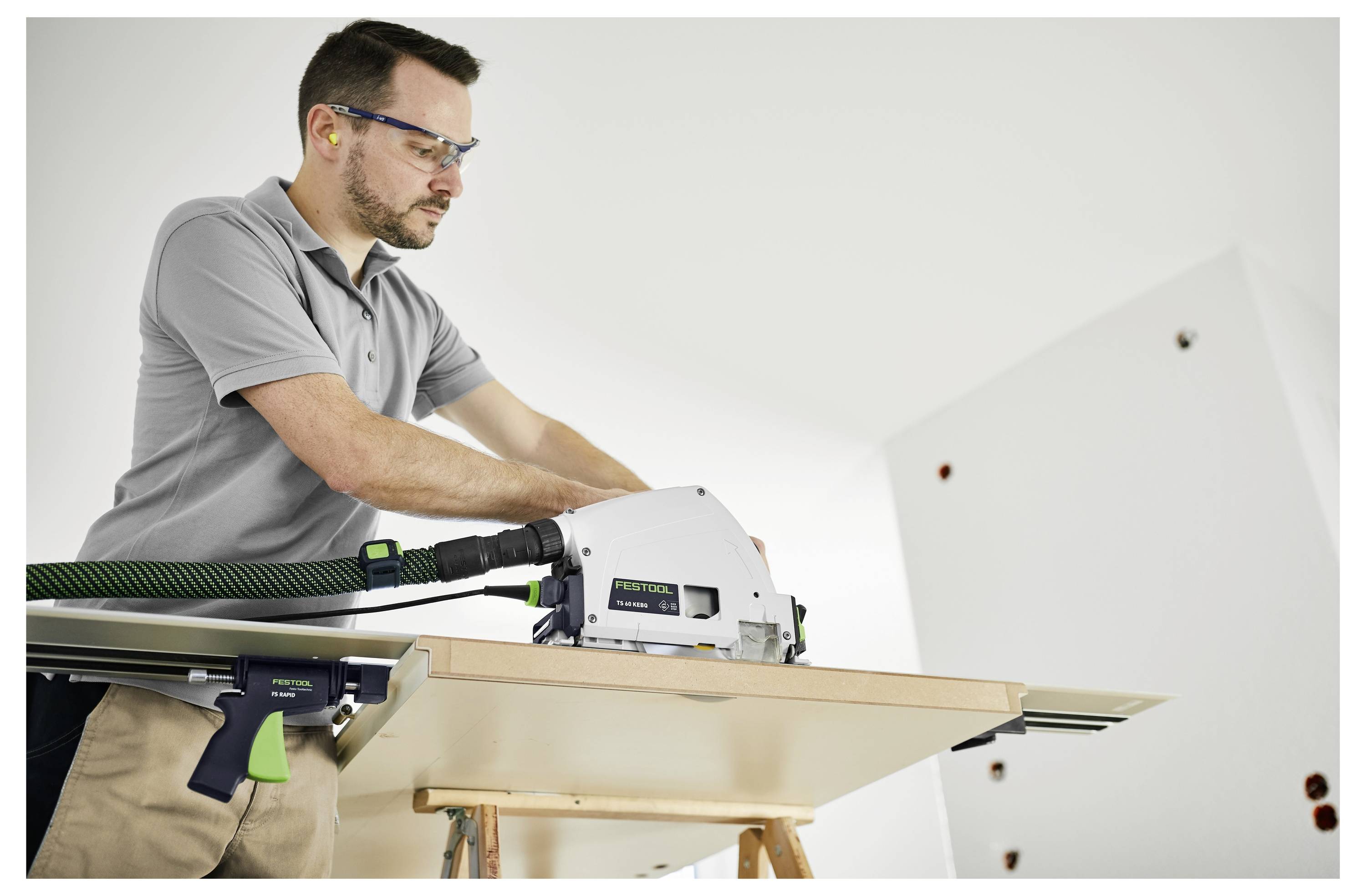 Muž používa motorovú pilu na rezanie drevenej dosky, pričom má nasadené bezpečnostné okuliare a ochranu sluchu v dielňovom prostredí.