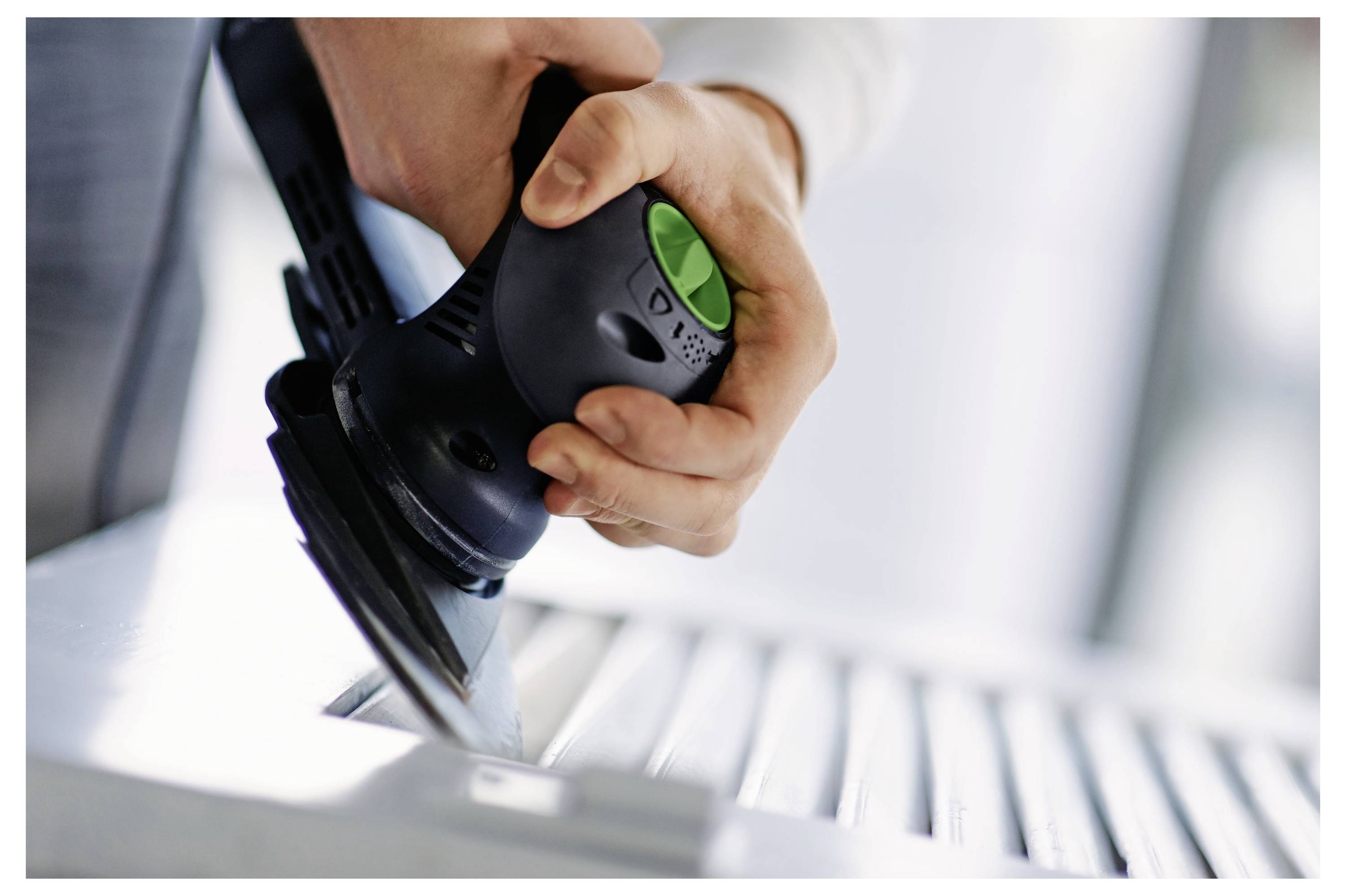 Osoba používa elektrickú brúsku na vyhladenie povrchu dreveného predmetu, pričom sa zameriava na ľavú stranu. Nástroj má zelené tlačidlo.