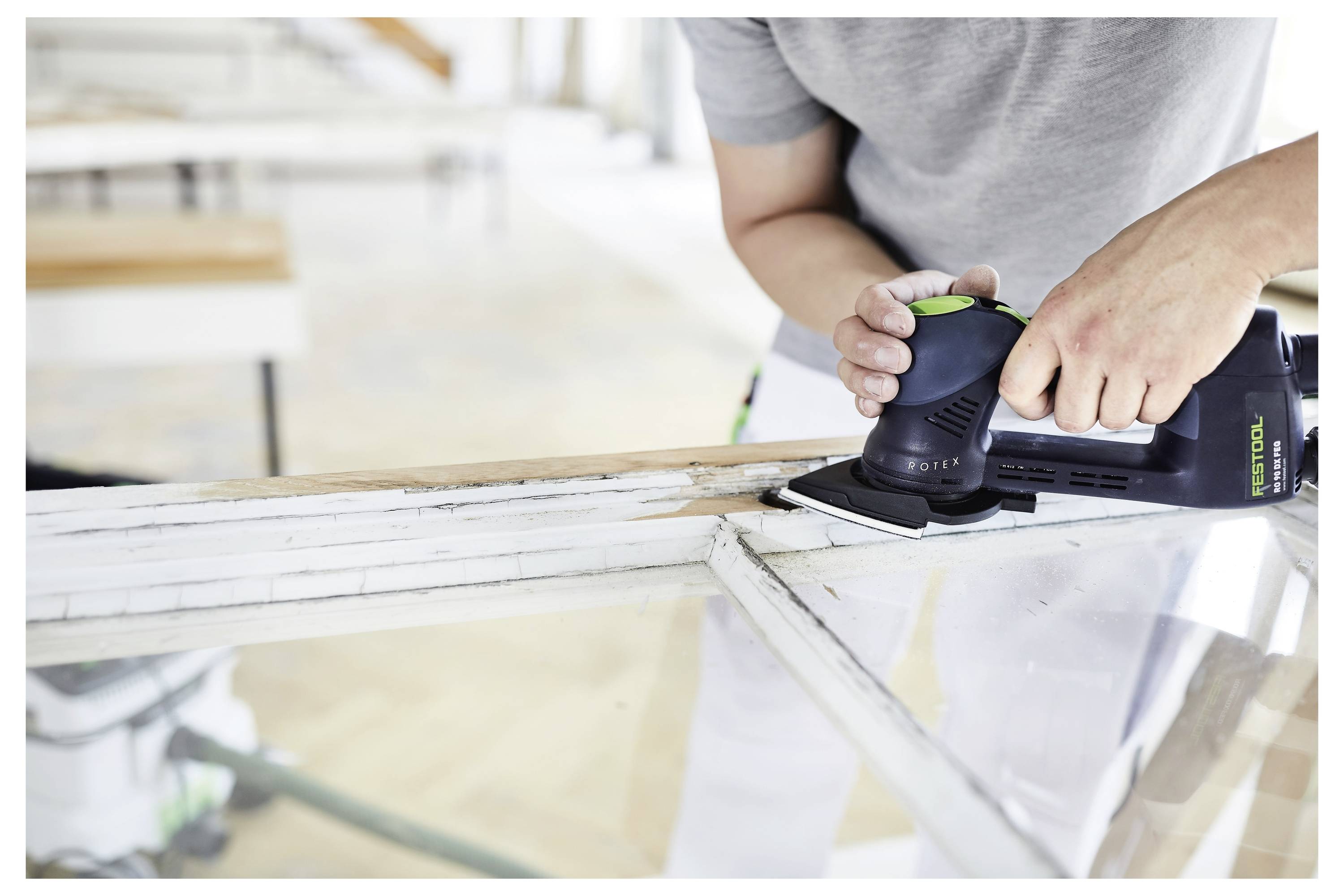 Osoba brúsi starý rám staného okna pomocou ručnej elektrickej brúsky, pričom sa zameriava na vyhladenie povrchu dreva v pracovnej dielni.