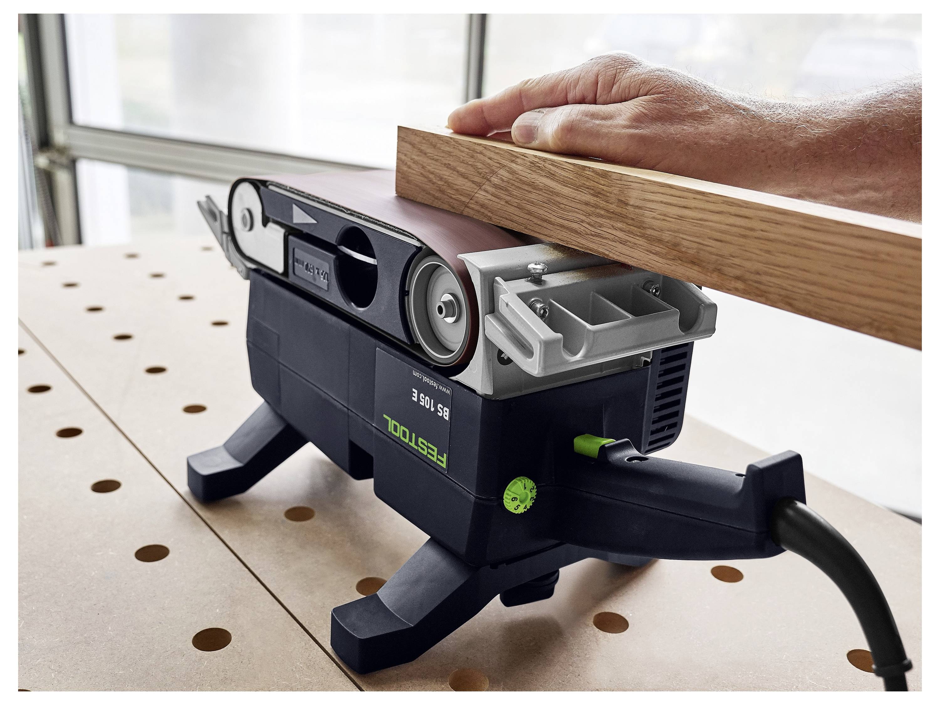 Osoba používa pásovú brúsku na vyhladenie drevenej dosky na perforovanej pracovnej doske v jasne osvetlenej dielni.