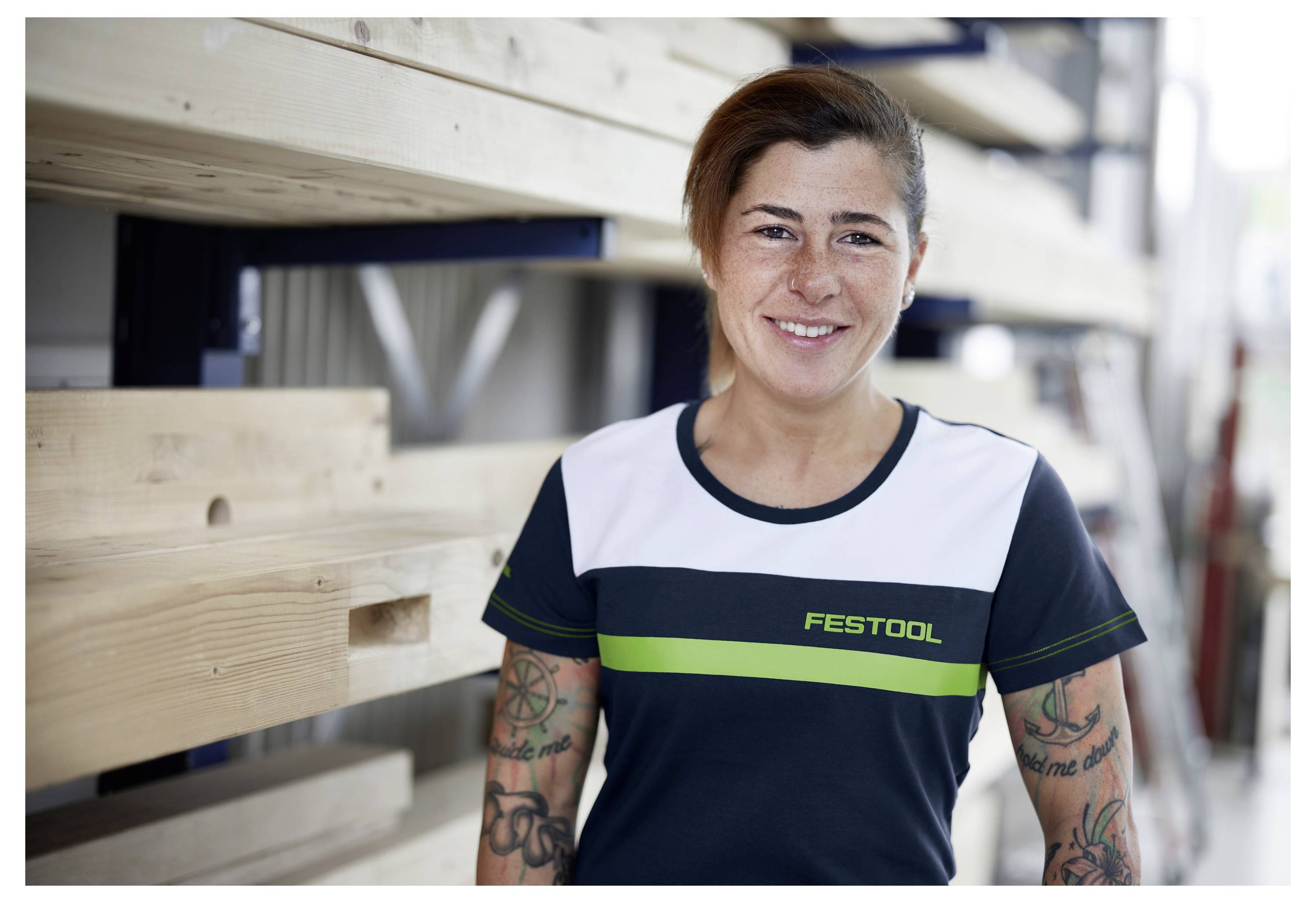 Osoba sa usmieva v dielni a má na sebe tričko s logom značky "Festool". V pozadí sú police s drevenými doskami.