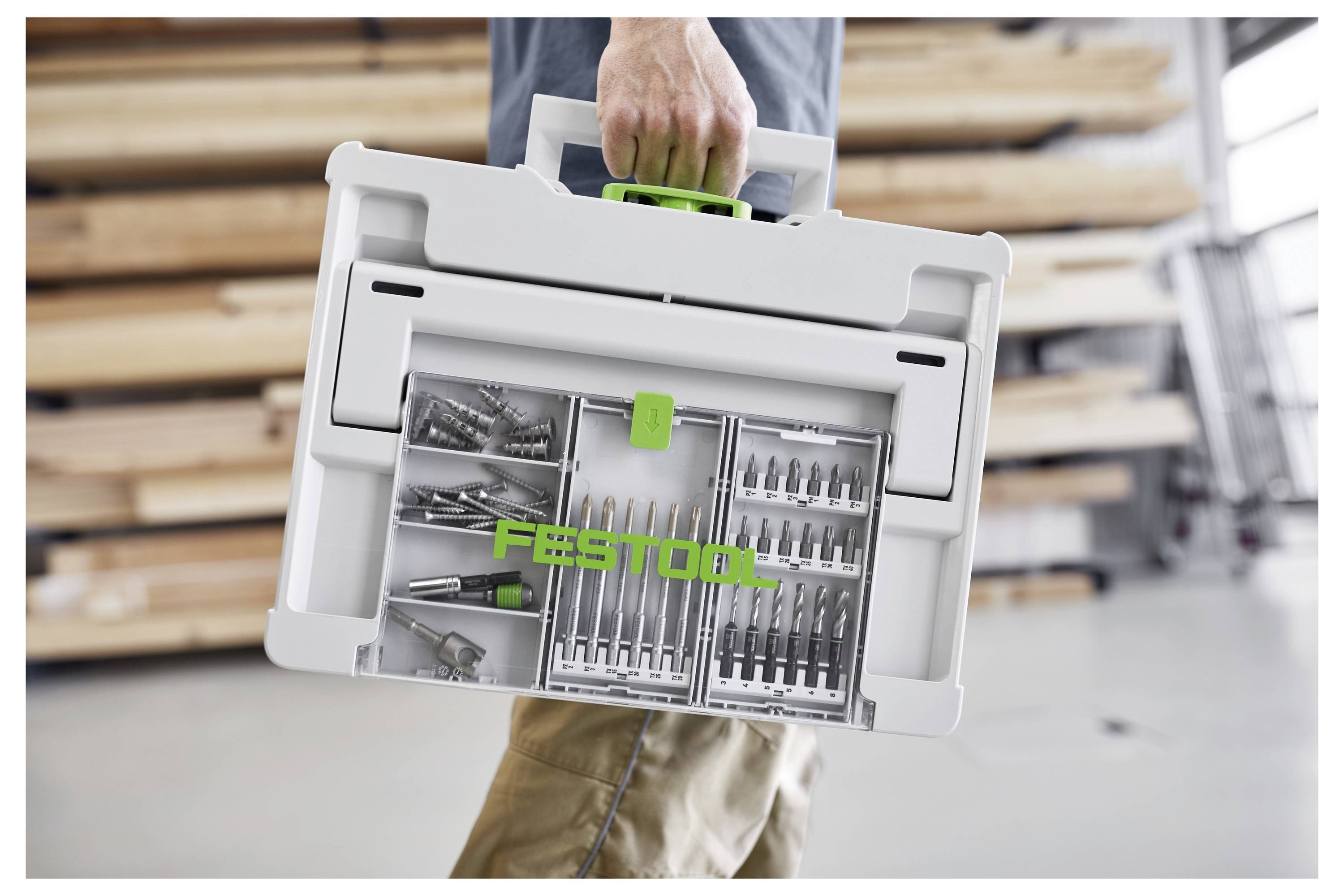 Osoba nesie Festoolov kufrík s rôznymi vrtákmi, na pozadí sú uložené drevené dosky a dielňové náradie.