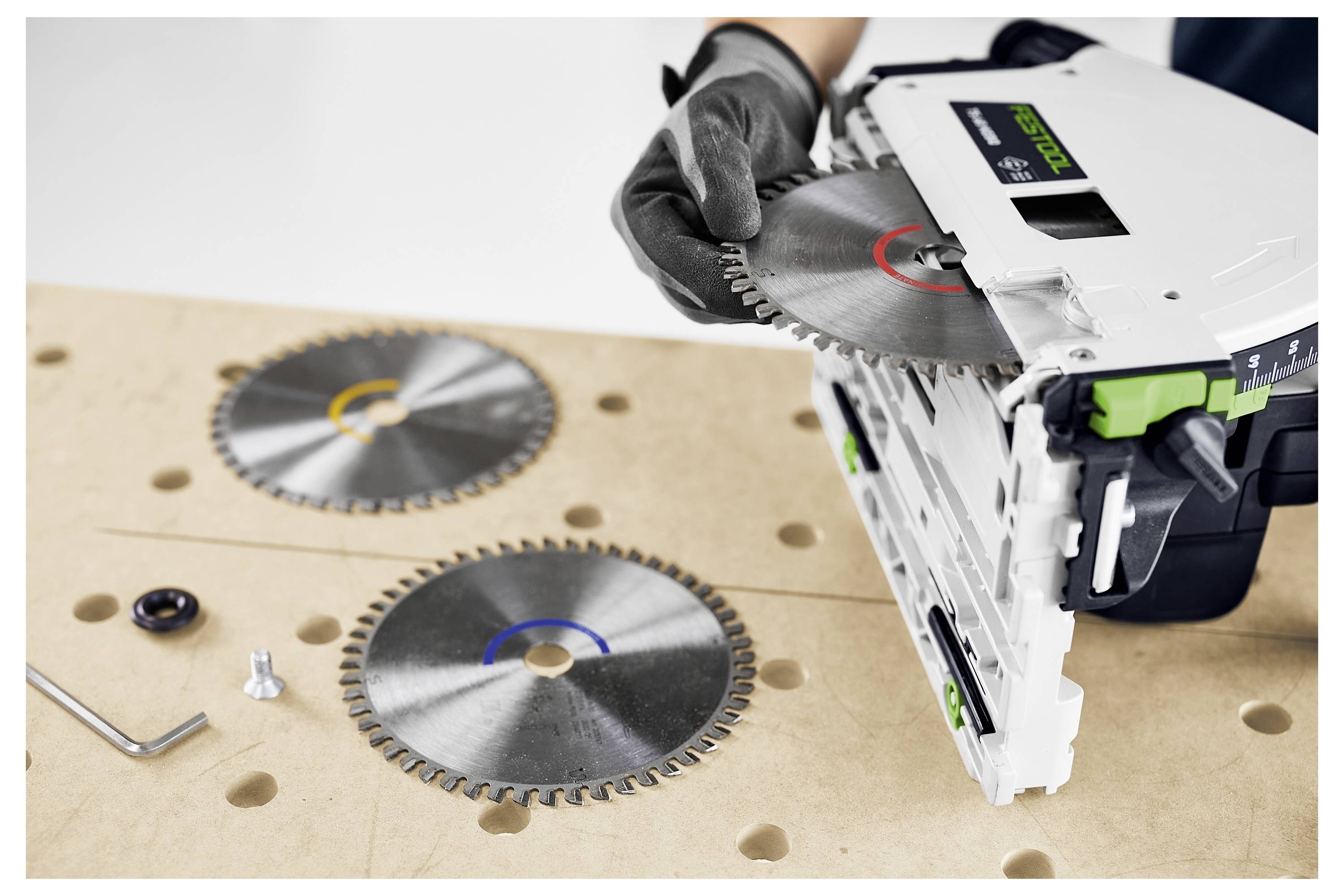 Osoba si mení čepeľ okružnej píly na pracovnom stole, pričom na povrchu sú viditeľné ďalšie dve čepele a niekoľko malých nástrojov.