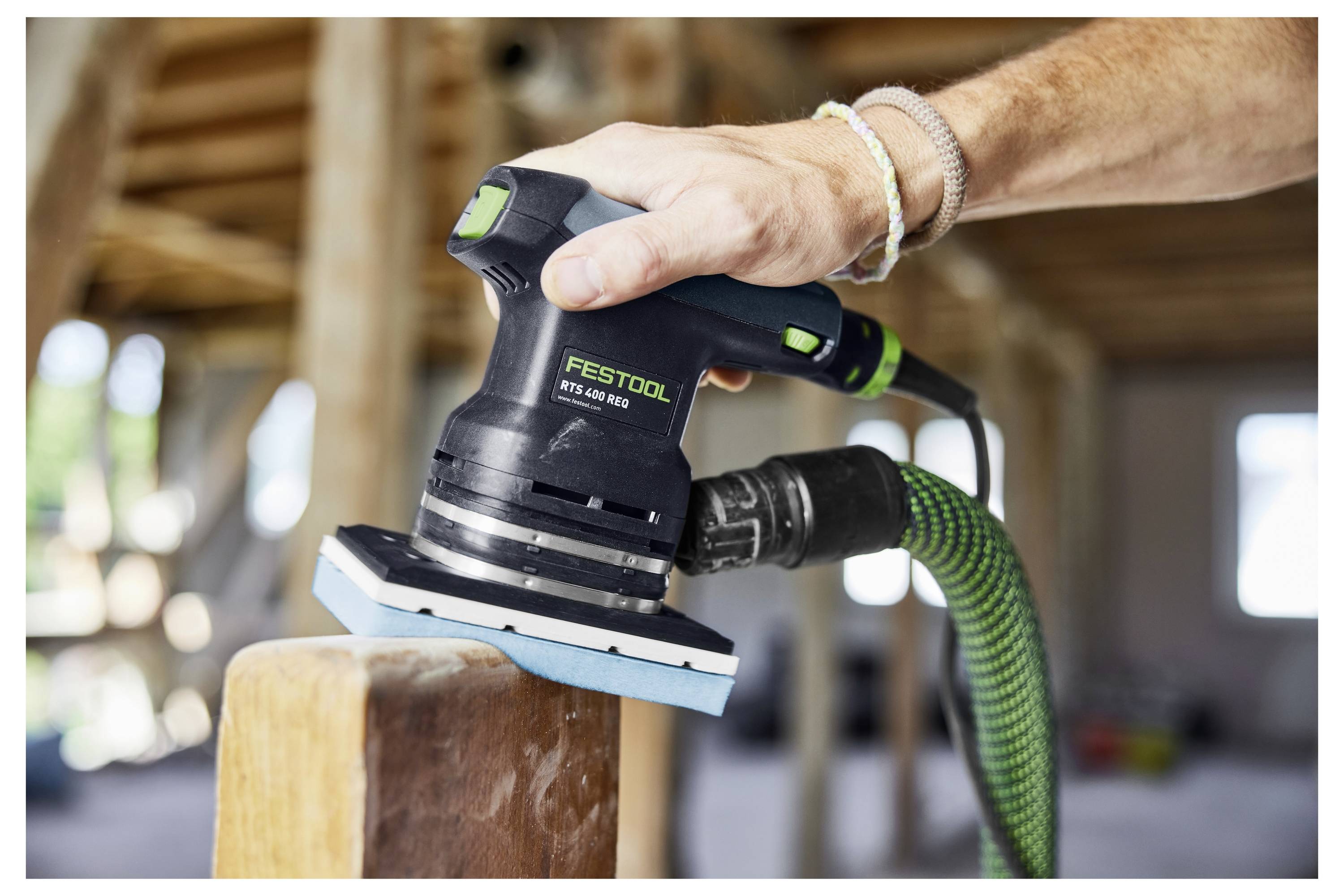 Ruka používa elektrickú brúsku Festool na drevenom nosníku v stavebnom prostredí. V pozadí sa svetlo filtruje cez okná.