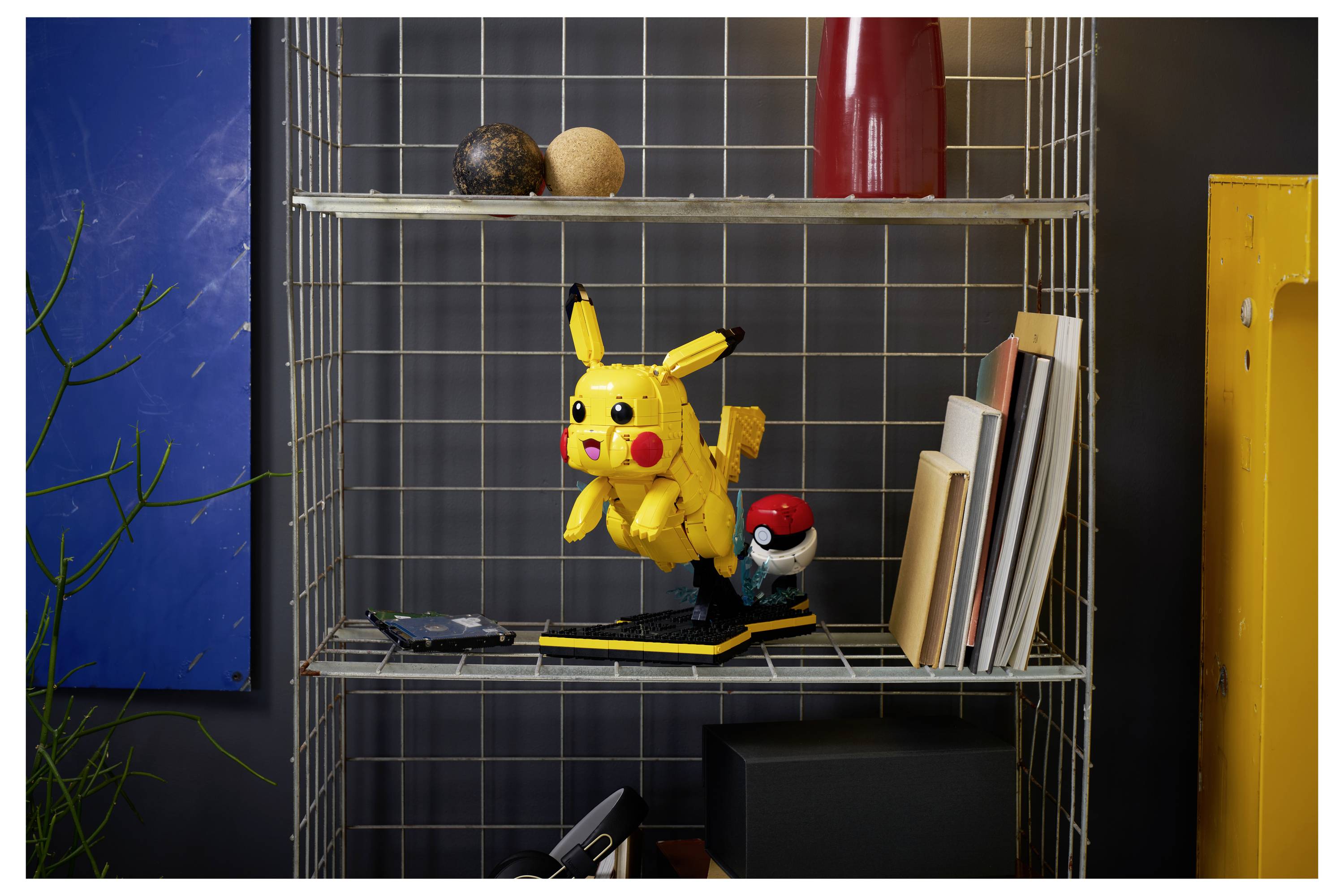 Polica s hračkou Pikachu a Poké Ball medzi rôznymi knihami a dekoračnými predmetmi, postavenou pred stenou z drôtenej siete.