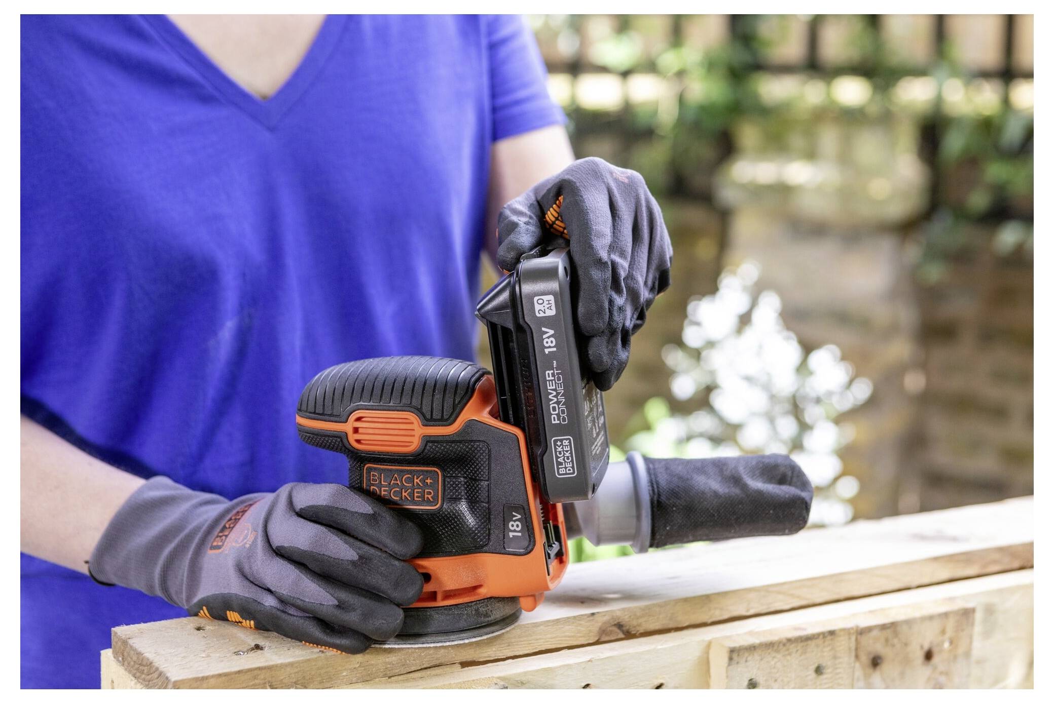 Osoba nosená rukavice používa bezdrôtovú brúsku Black+Decker na drevenom povrchu. Pozadie ukazuje vonkajšie prostredie so zelenňou.