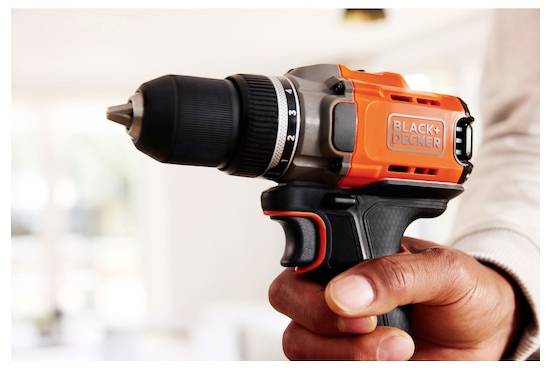 Osoba drží oranžovo-čiernu elektrickú vŕtačku značky „Black+Decker"; pozadie je jemne osvetlená miestnosť.