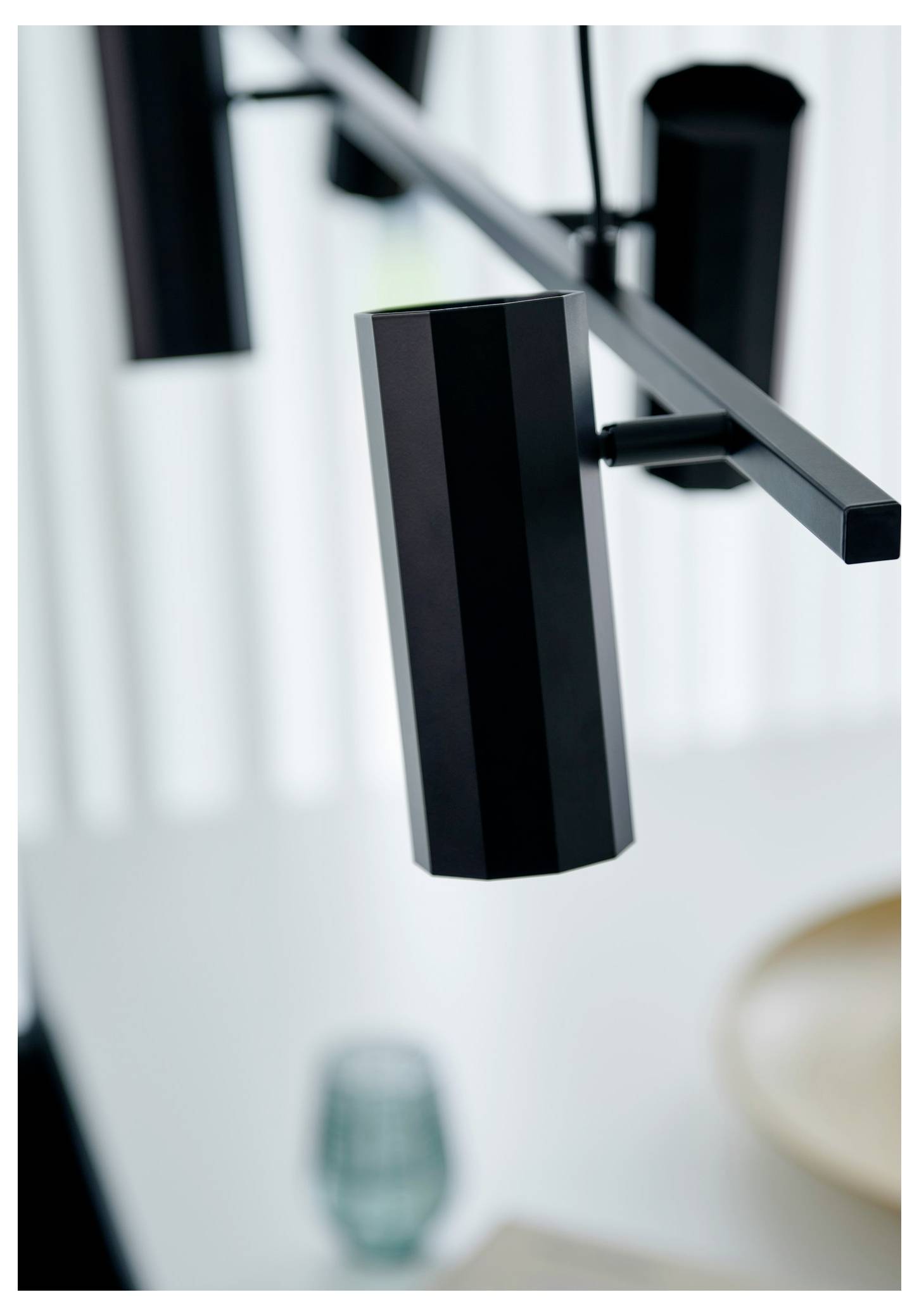 Zblízka moderný čierny stropný svetelný prvok s geometrickým dizajnom, umiestnený nad rozmazaným prostieraním jedálneho stola.