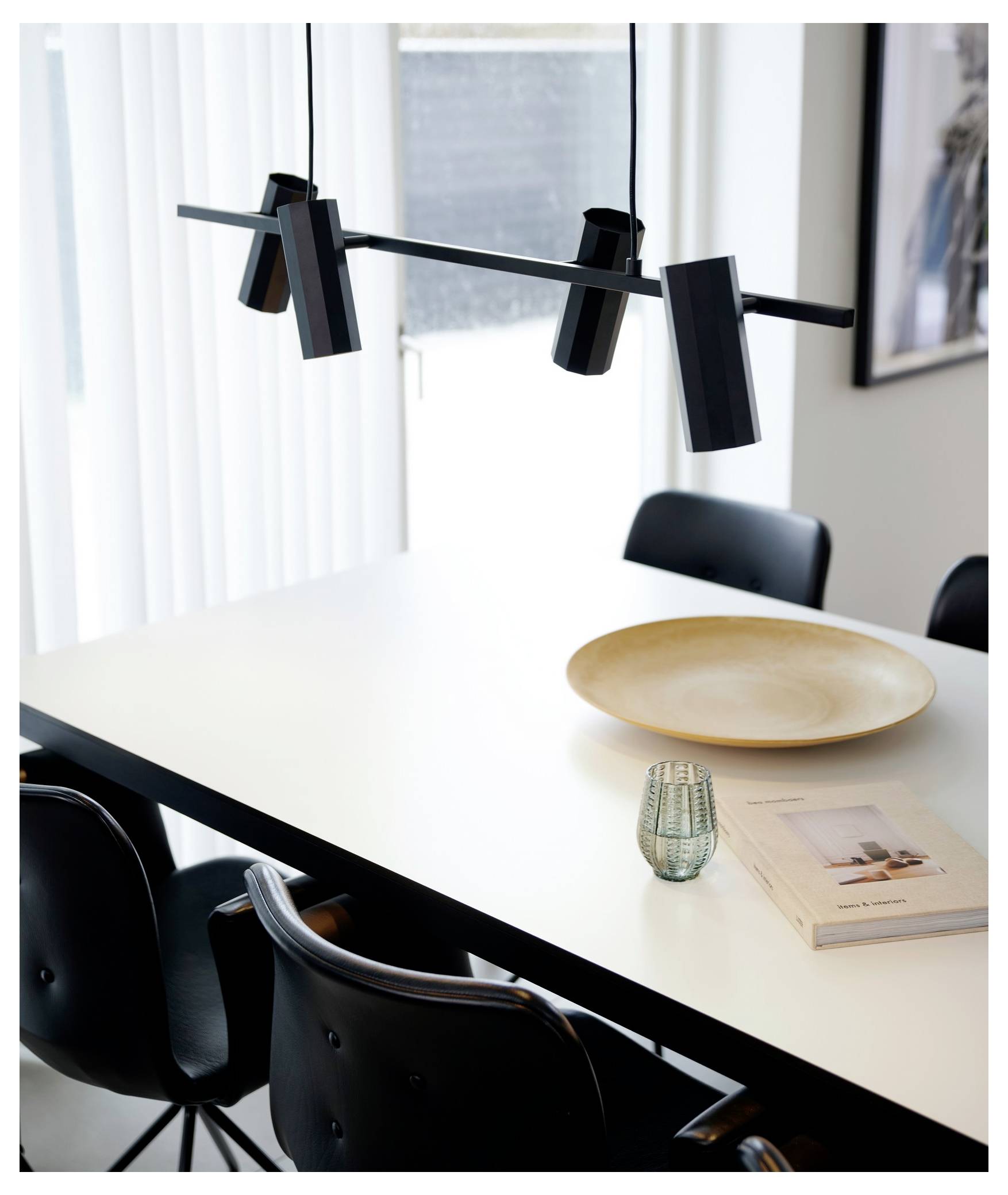 Moderná jedálenská oblasť s bielym stolom, čiernymi stoličkami, čiernym stropným svetelným teliesom, keramickým tanierikom, malou vázou a dvomi knihami.