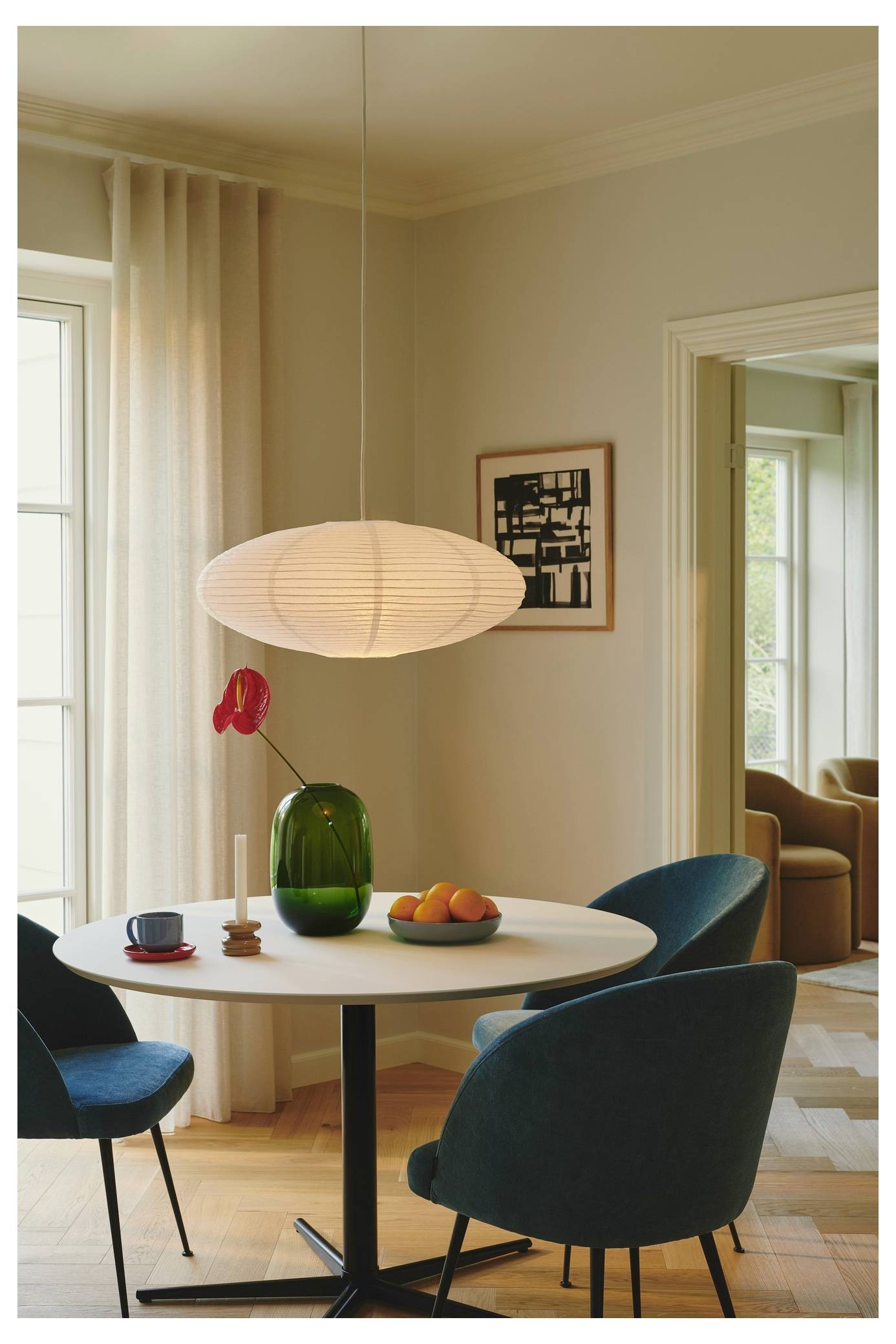 Okrúhly jedálenský stôl s tmavě modrými stoličkami, zelená váza s červeným kvetom, miska s pomarančmi a visiaca biela lampa v útulnej miestnosti.