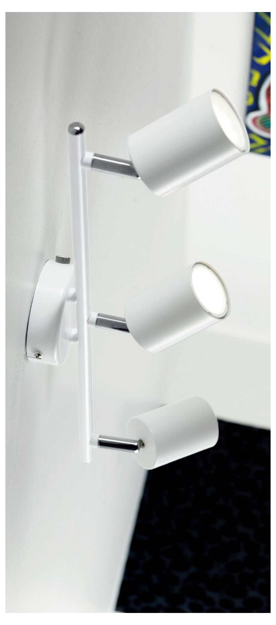 Trojitá nástenná bodová lampa v bielej farbe s chrómovým držiakom, umiestnená v kútiku miestnosti s pohľadom na farebné umelecké dielo.