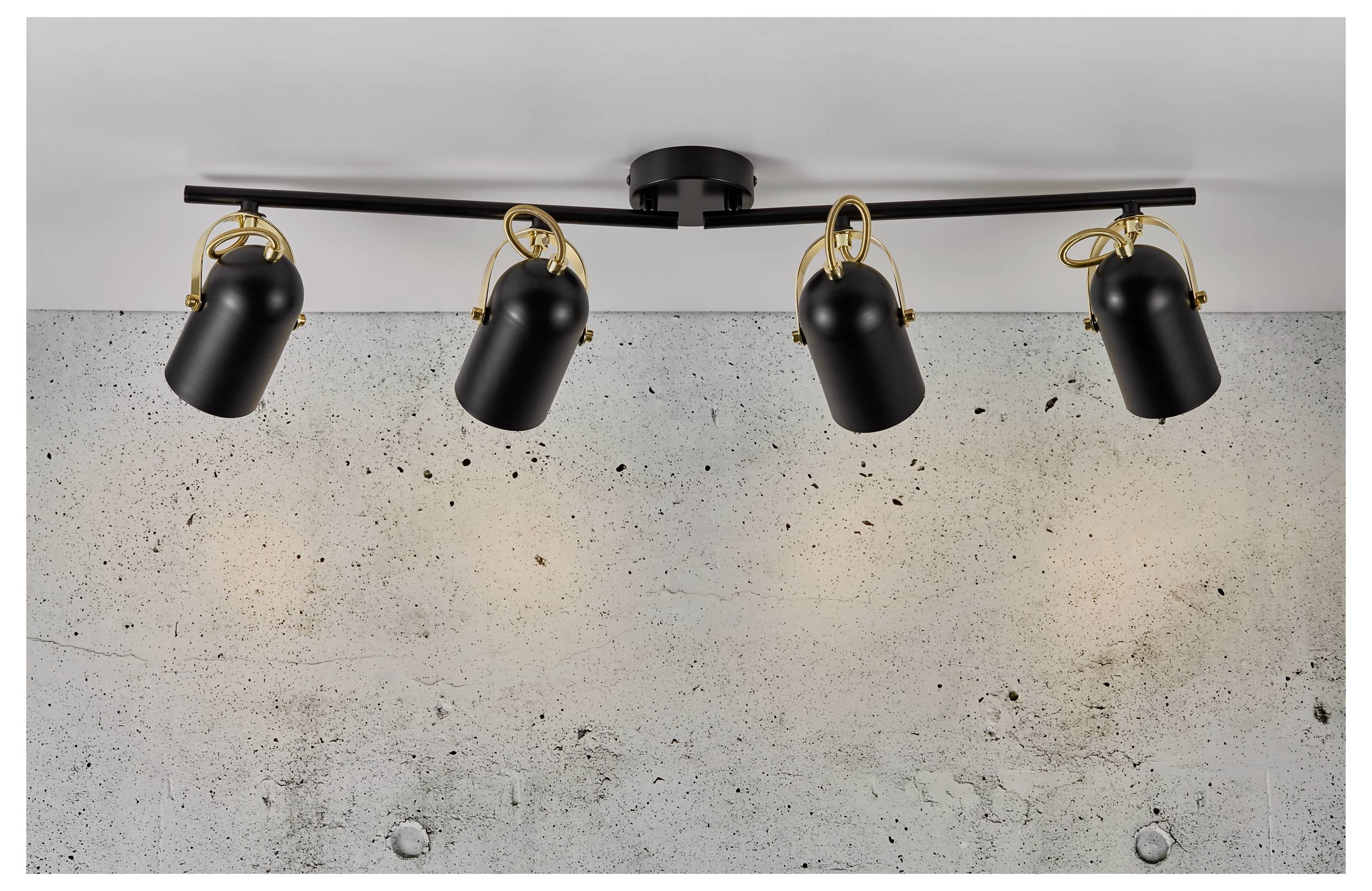 Stropný svetelný prvok so štyrmi nastaviteľnými čiernymi reflektormi, namontovaný na čiernej tyči s dekoratívnymi zlatými krúžkami, na pozadí betónovej steny.