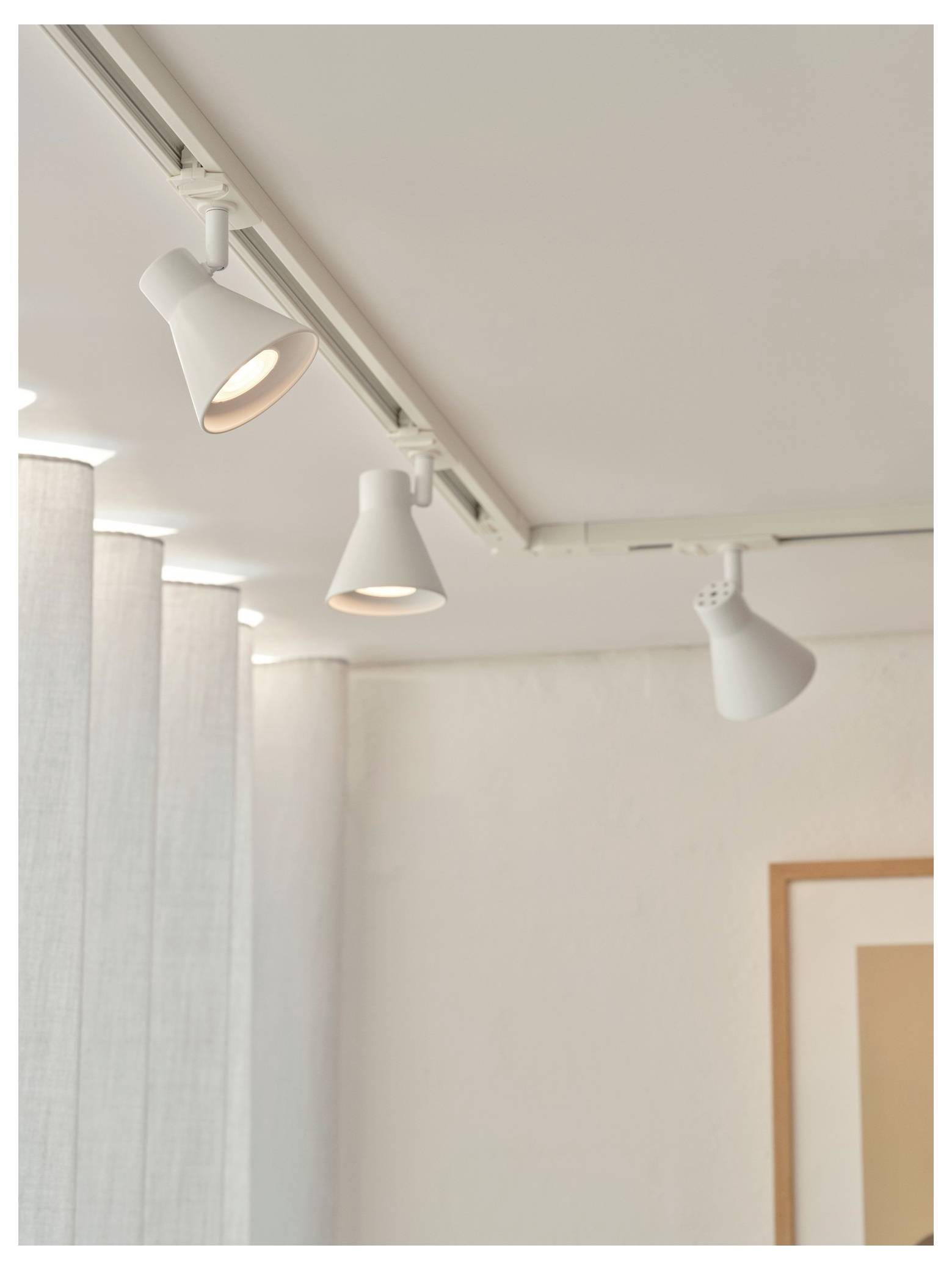 Stropný koľajnicový systém s tromi bielymi nastaviteľnými reflektormi osvetľujúcimi modernú, minimalistickú interiérovú priestor s bielymi stenami a zarámovaným obrázkom.