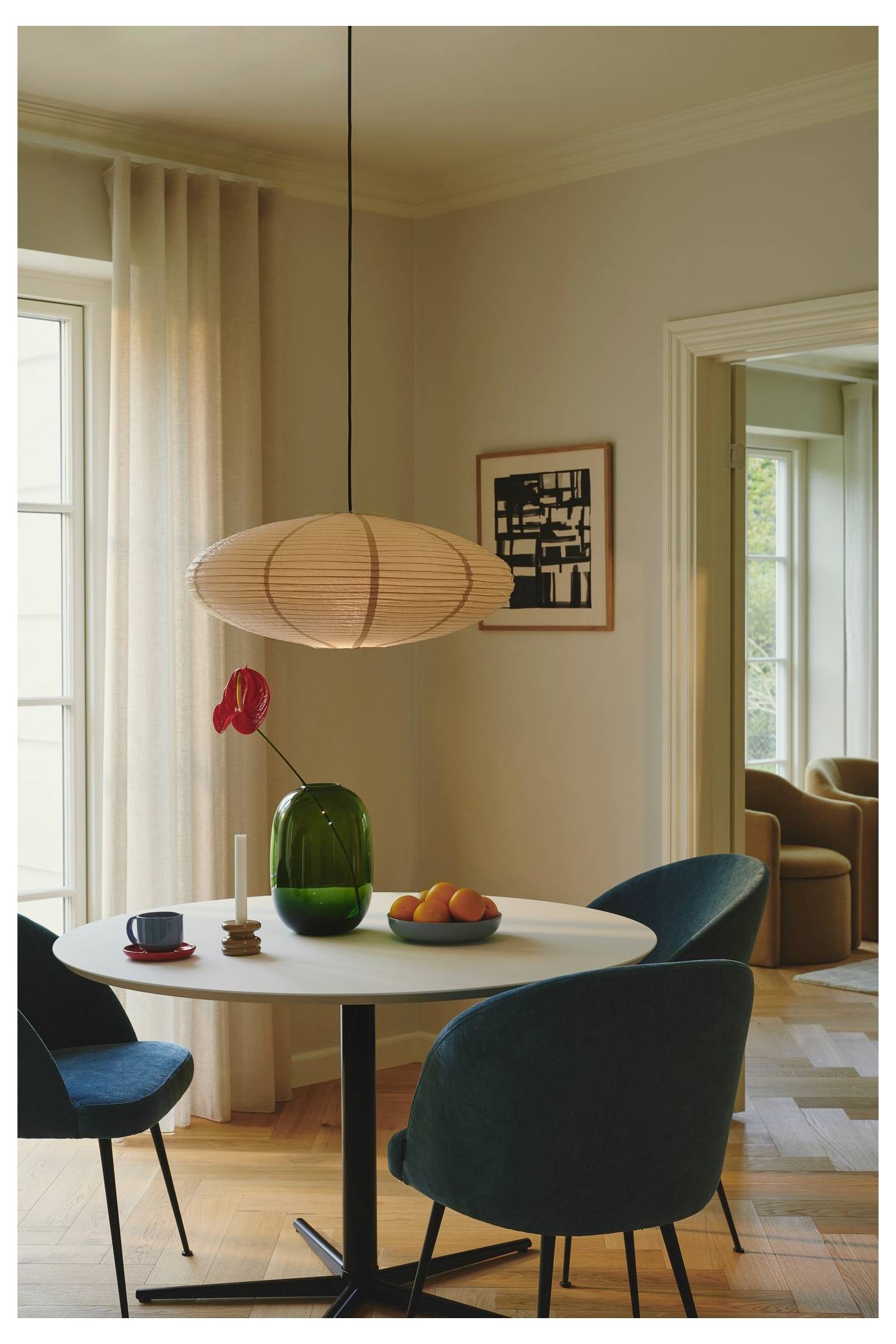 Moderná jedáleň s okrúhlym bielym stolom, tyrkysovými stoličkami, zelenou vázou s červeným kvetom, mísou s ovocím a závesným svetlom; slnečné svetlo sa pretláča cez priehľadné záclony.