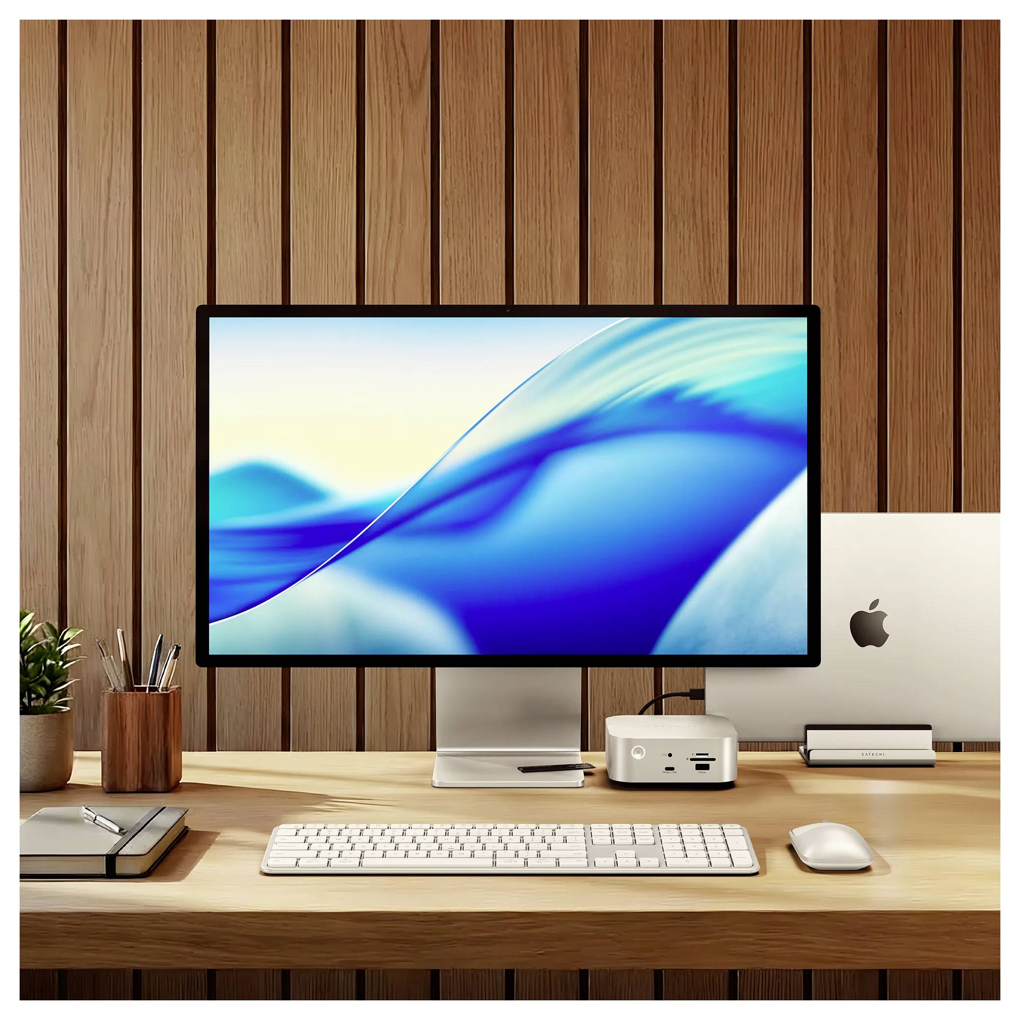Moderný pracovný stôl s veľkým monitorom zobrazujúcim modré abstraktné vlny, počítač Apple, klávesnica, myš a príslušenstvo na pozadí drevosteňovej steny.