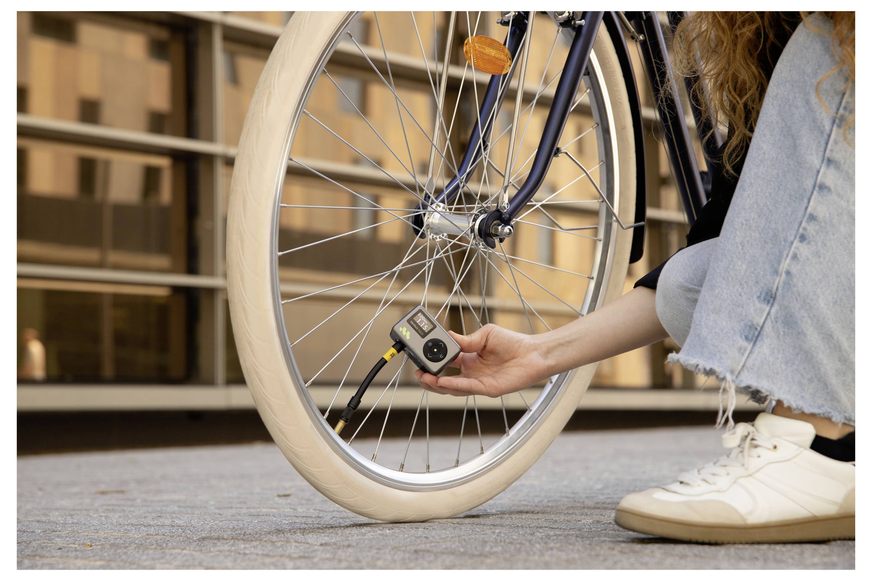 Osoba v rifliach a tenisky používa tlakomer na kontrolu pneumatiky bicykla na spevnenej ploche pred budovou.