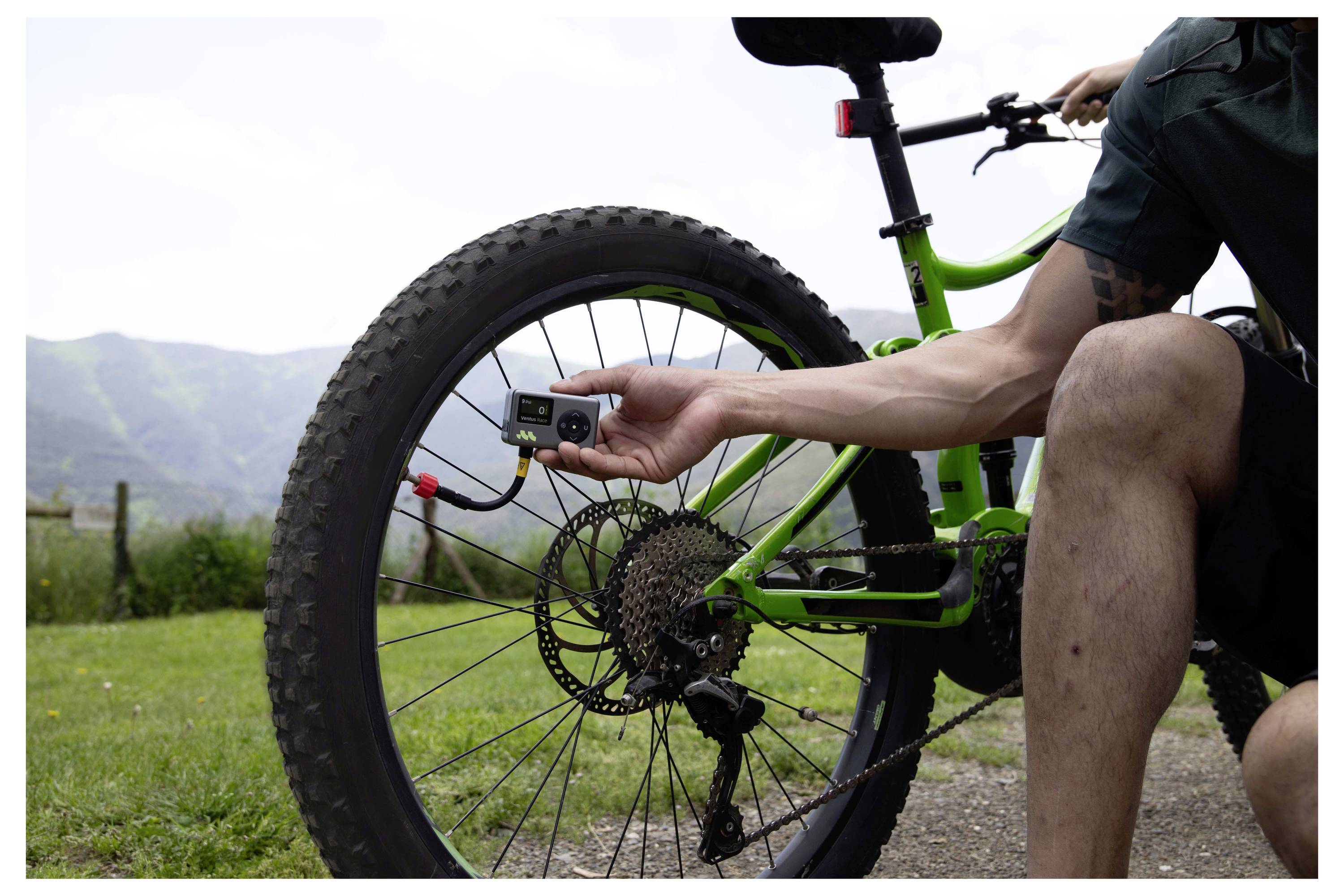 Osoba na zelenom horskom bicykli kontroluje tlak v pneumatike zadného kolesa pomocou digitálneho manometra vonku na trávnatej ceste s kopcami v pozadí.